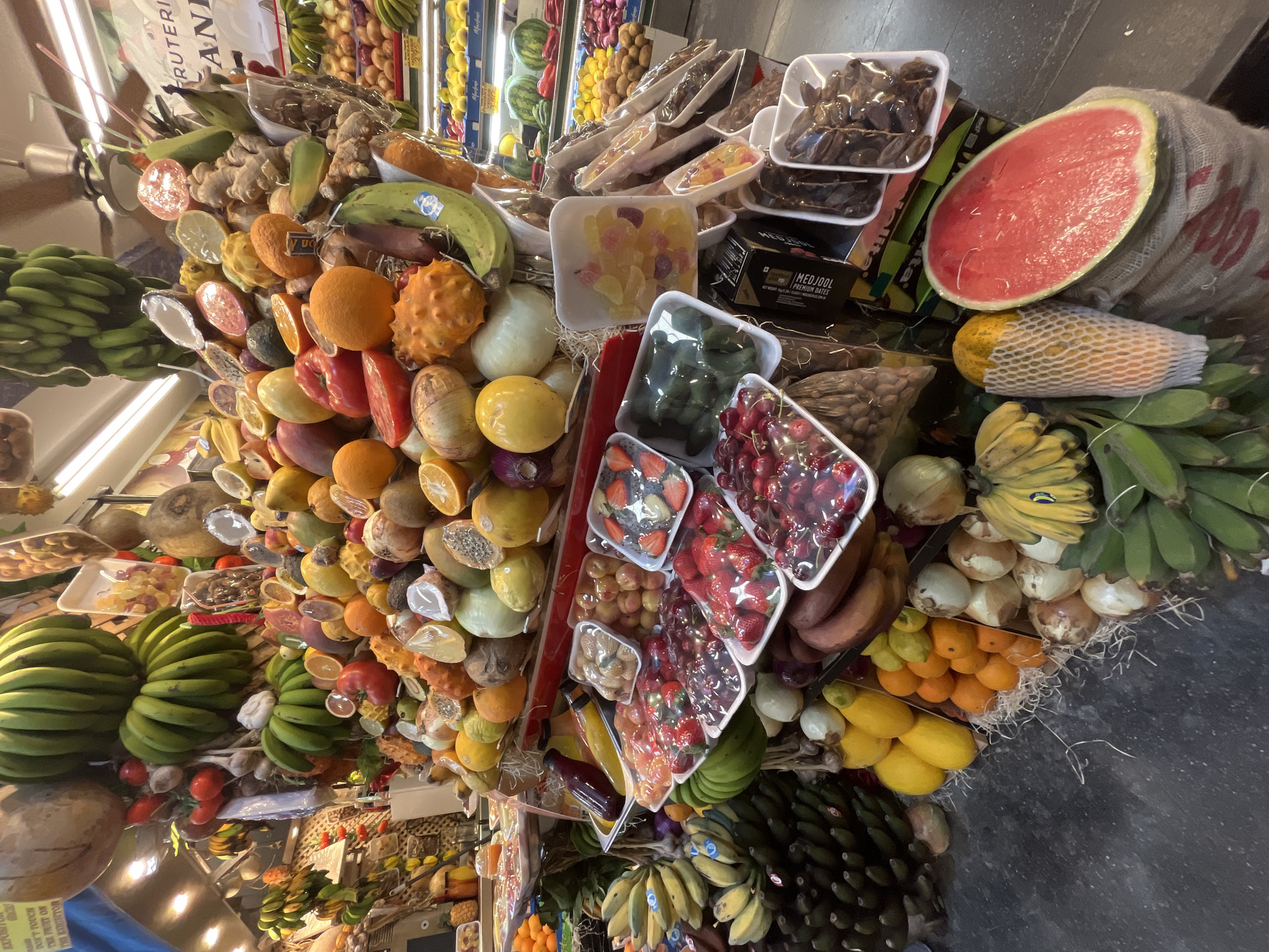 The image shows a fruit and vegetable stand, likely inside a market in Las Palmas, Spain, stacked high with various items. There are fruits like bananas, watermelons, oranges, and other citrus fruits. Also present are packaged goods such as dates and strawberries. The scene is densely packed with goods, offering a colorful display.
