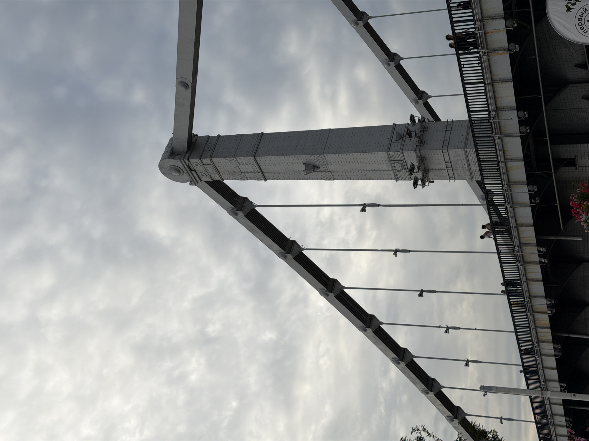 The image shows a suspension bridge in Moscow, Russia, under a cloudy sky. The bridge's large, grey metal supports are prominent, with cables extending to the deck. Several people are visible walking on the pedestrian walkway of the bridge, some appearing to be tourists or locals enjoying the view. The bridge structure is detailed, with rivets and decorative elements visible on the supports. Below the walkway, the underside of the bridge shows complex engineering with visible beams and supports. In the foreground, some pink flowers are partially visible, suggesting a park or green space adjacent to the bridge. The overall atmosphere is overcast, with diffused light from the sky.
