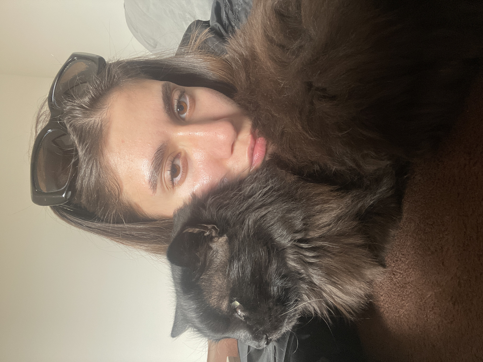 This indoor portrait captures a young person and a cat in close proximity. The person, appearing to be a woman, has long dark brown hair and light brown eyes. She is seen from the chest up, with sunglasses pushed onto the top of her head, one arm of which might have the faint, partial text "VEGA" visible. Her skin has a natural sheen, and her lips are slightly parted. She is wearing a dark, possibly black, jacket or top.
Cuddled next to her is a large, long-haired cat with dark, potentially black or very dark grey, fluffy fur. Its face is close to the person's, and one of its eyes, partially visible, has a light glint. Long whiskers are also visible. The cat appears calm, nestled comfortably against the person.
The setting is indoors, likely a home environment, with a plain, light-colored wall in the upper background and a textured brown carpeted surface in the lower right. The lighting is soft, creating highlights on the person's skin and the cat's fur. The overall impression is one of affectionate companionship. No other discernible text is visible in the image. The location context for this scene is Sofia, Bulgaria.