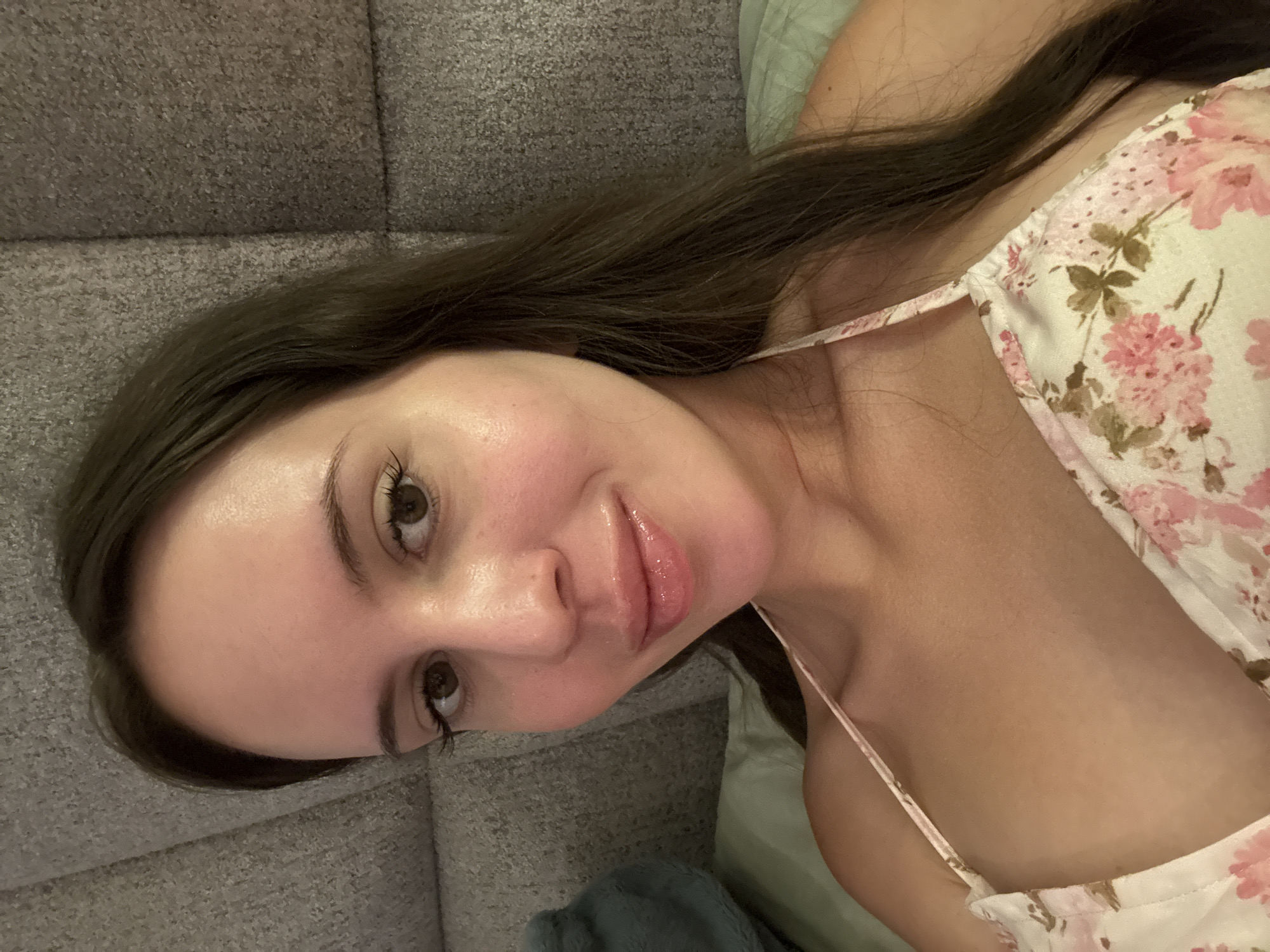 A woman with long, dark brown hair lies on her side, looking at the camera. She is wearing a floral-patterned top with thin straps. Her skin appears dewy, and her lips are glossy. She is propped up against a textured, grey headboard or wall. The lighting is soft, suggesting an indoor setting, likely a bedroom. There is a hint of a blue pillow or blanket visible on the left side of the frame.