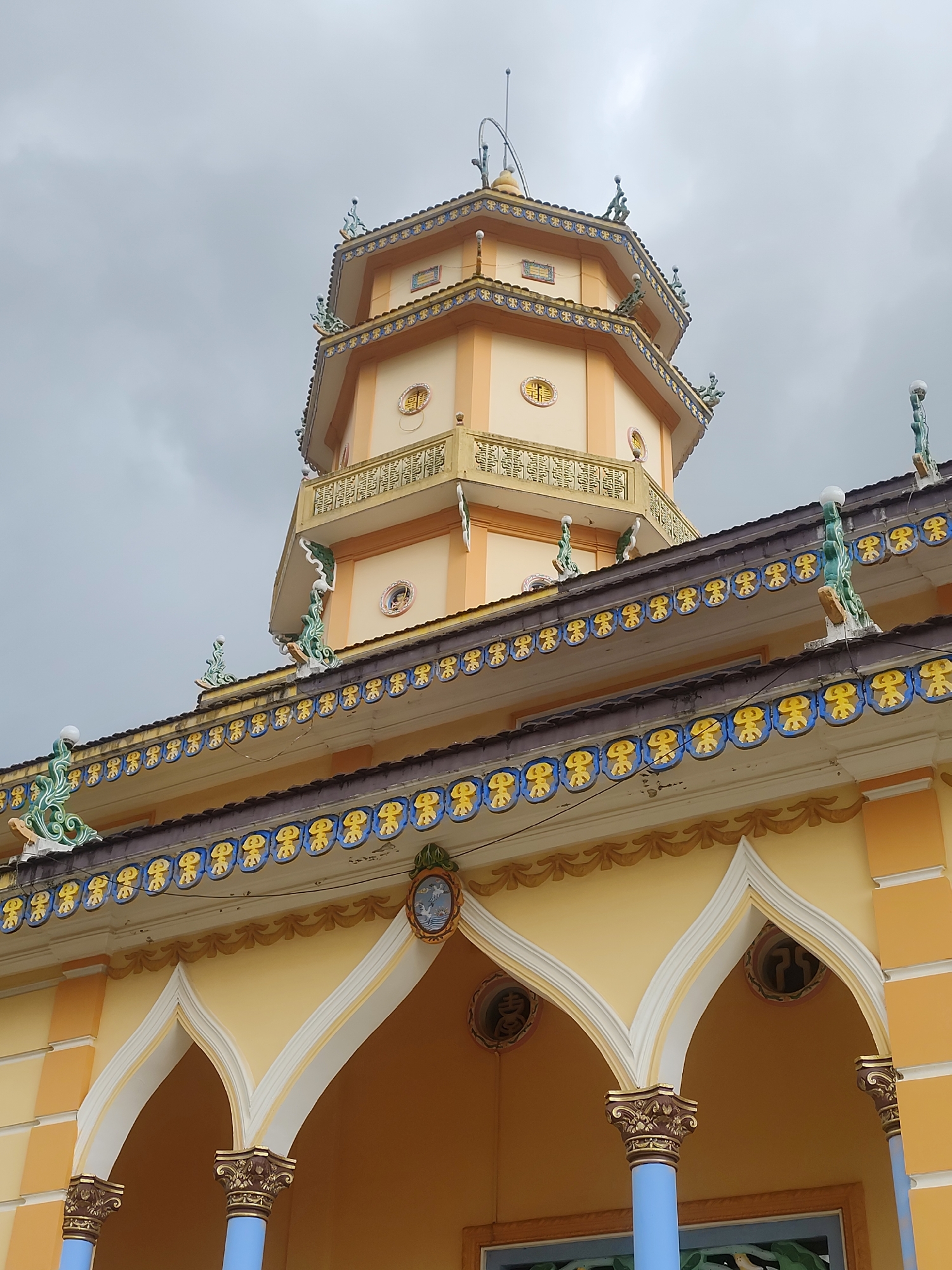 The image shows a close-up of the facade and tower of a Buddhist temple in Lộc Quý, Vietnam. The temple features traditional Vietnamese architecture with yellow and orange painted walls, intricate decorative moldings along the rooflines, and octagonal tower with multiple tiers. The tower is topped with a finial and has decorative elements on each level. The facade of the temple is adorned with ornate arches supported by columns, and decorative circular motifs are visible. The sky is overcast with gray clouds.