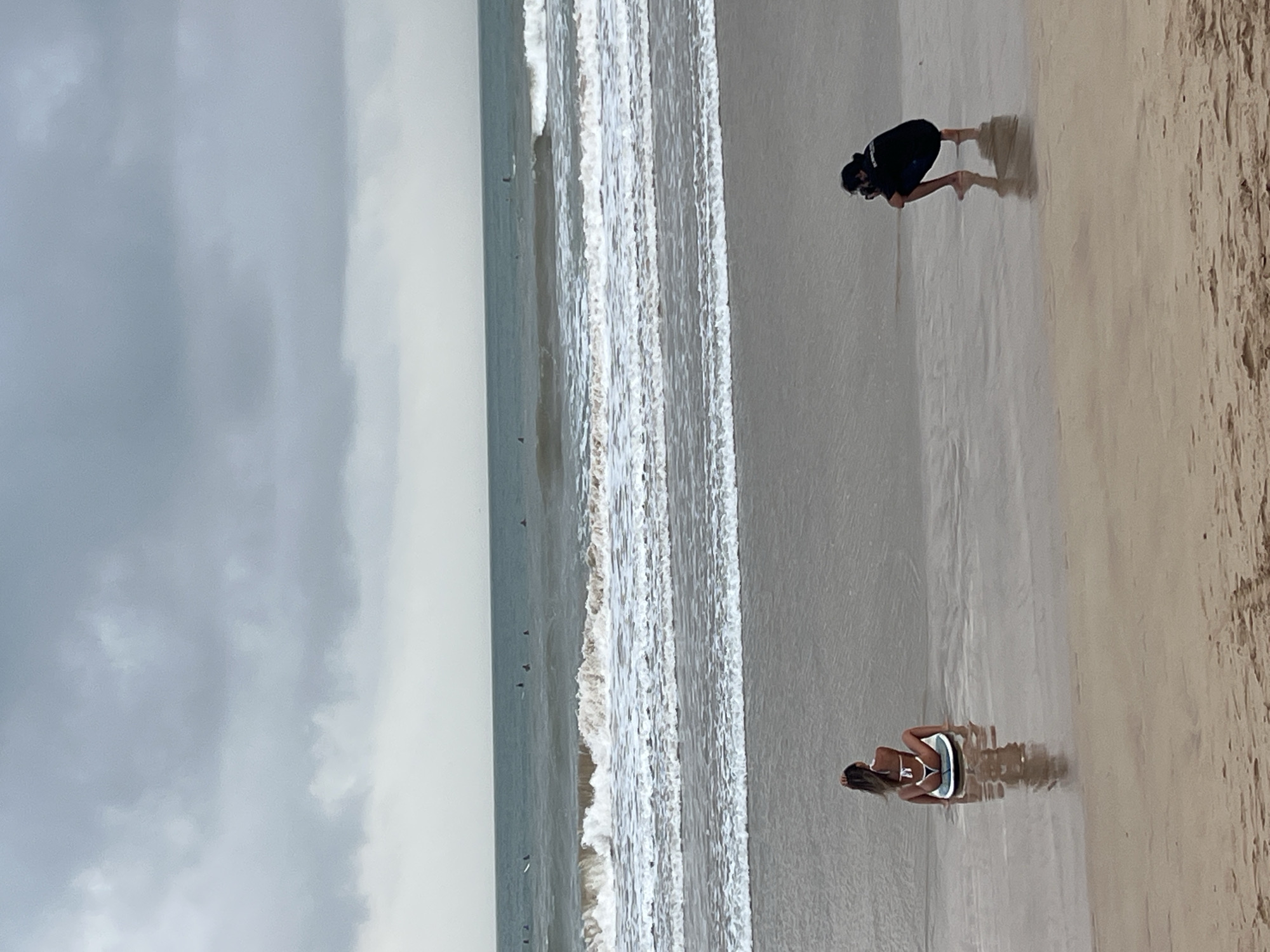 In Peleana, Sri Lanka, on a beach with gray, cloudy skies, two people are present. A woman in a white bikini sits on a surfboard in the shallow water, facing away from the camera, with her hand in her hair. To her right and further back, a person wearing a black shirt and dark shorts crouches down, appearing to take a photograph of the woman with a camera. The ocean is calm with small waves breaking, and several surfers are visible in the distance. The sand in the foreground is wet and reflective from the water.