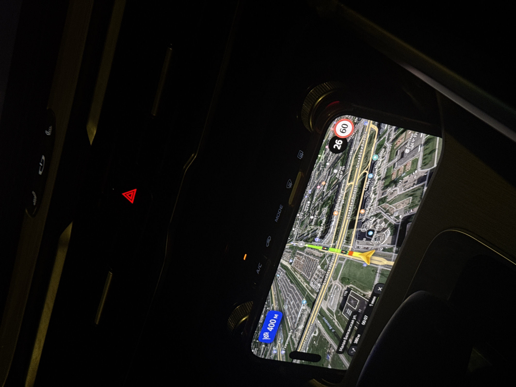 The image shows the interior of a car at night, with the car's navigation system prominently displayed on a screen. The navigation map shows a route with a destination of "400 m" and a current speed of "26" km/h, with a speed limit indicated as "60" km/h. The map is oriented over an urban area, specifically identified by street names and landmarks visible on the screen. These include "Малая Балканская ул." (Malaya Balkanskaya Street), "р. Волковка" (Volkovka River), and other Russian street names and place names like "Дунайский проспект" (Dunayskiy Prospekt). The navigation system appears to be in Saint Petersburg, Russia, based on the presence of these street names. The car's interior is dark, with only the glow from the navigation screen and the red illumination of the hazard light button providing light.