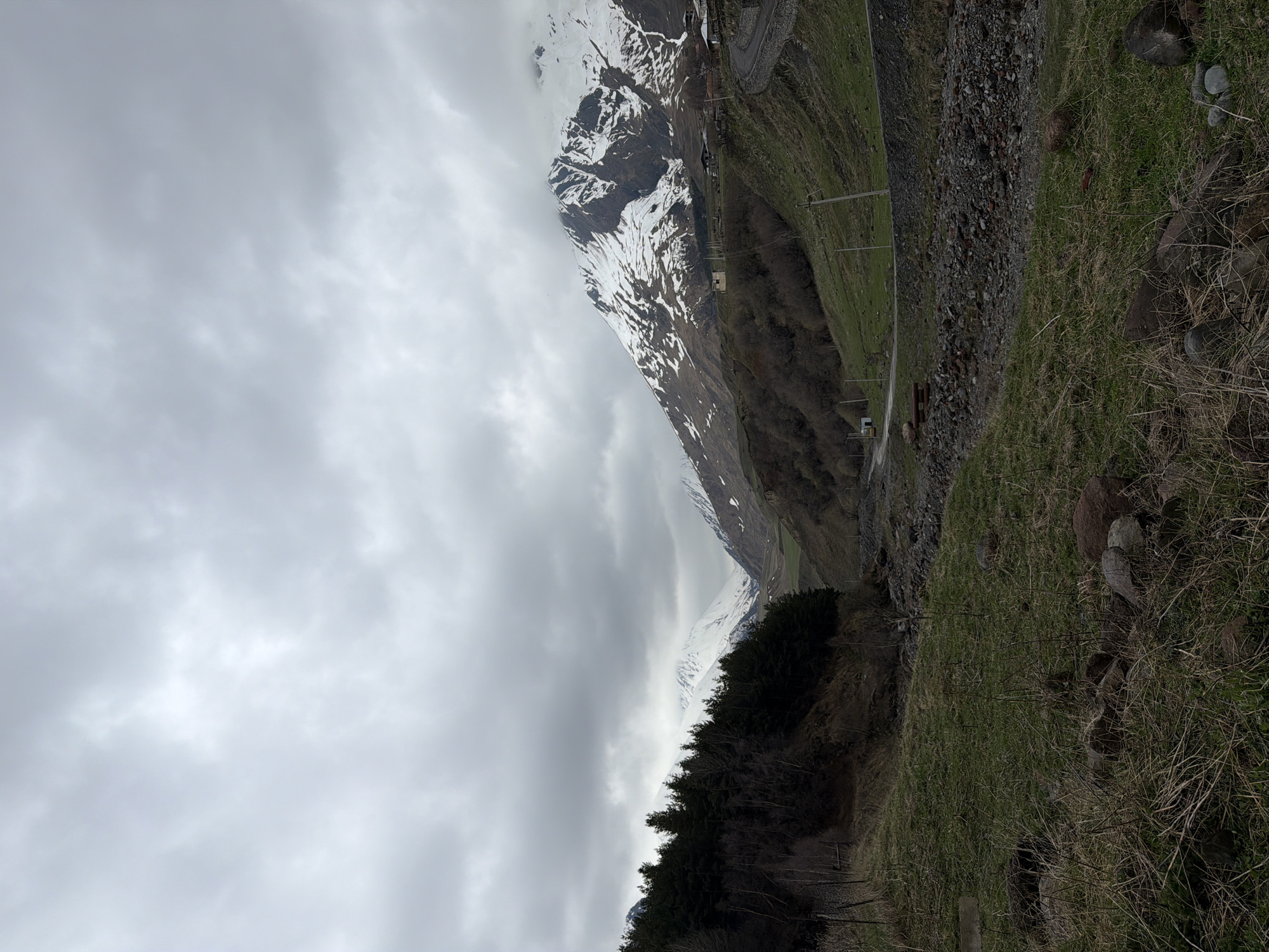 The image shows a mountainous landscape in Khurtisi, Georgia, under a cloudy sky. The left side of the image is dominated by gray clouds. On the right, a steep mountain slope descends, covered in green grass and some bare patches of soil. A rugged, rocky area runs along the upper right edge. A winding road is visible on the mountain, with occasional utility poles and a small white structure. Further in the distance, snow-capped peaks are visible. In the foreground, there is a treeline.