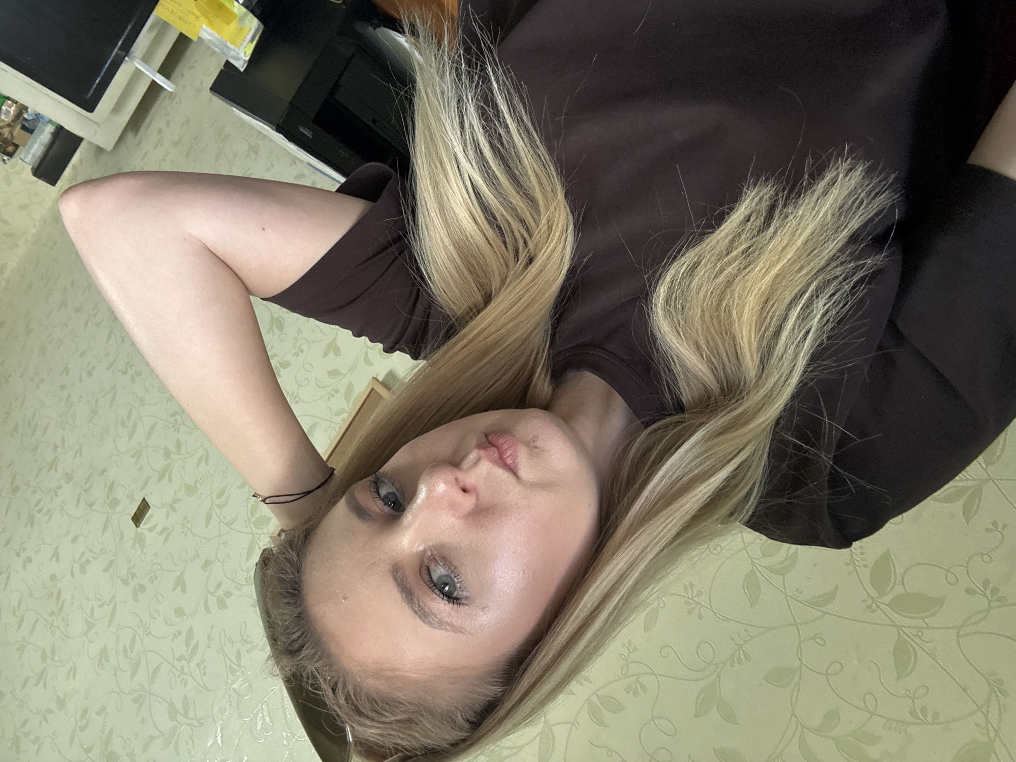 The media file is a close-up, low-angle, eye-level shot of a young woman lying down. She has fair skin, light blue eyes, and long blonde hair. Her hair is styled in loose waves and falls around her face and shoulders. She is wearing a dark brown t-shirt. Her left arm is bent, with her hand resting on the back of her head. She is looking directly at the camera with a neutral expression. The background is a light green wallpaper with a subtle floral pattern. There are some blurry objects in the background, including what appears to be a desk or shelf with electronic equipment. The lighting suggests an indoor setting, possibly during the daytime. No specific time of day, weather, or location cues are discernible beyond it being indoors. There is no visible text in the media file.