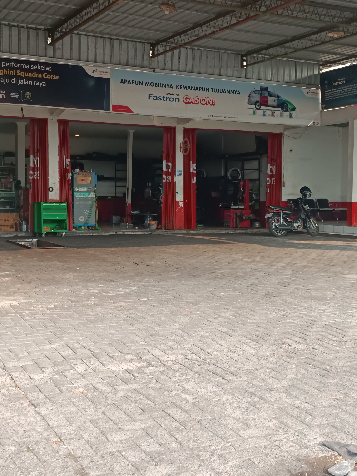 The image depicts the exterior of a Pertamina Fastron service station located on Jalan Raya Imogiri in Imogiri, Indonesia. The service station features red-and-white striped roll-up doors, with a large banner above displaying promotional material for Pertamina products, including "Fastron Gas On!" and a graphic of a car. Inside the service bay, visible through the open doors, are various pieces of automotive service equipment, including what appears to be a tire changer and oil dispensing units. A green tool chest sits outside one of the bays. A black motorcycle is parked on the paved area in front of the service station, and seating is visible in the background. The ground is paved with interlocking bricks, and the overall scene suggests a functional automotive service facility operating in daylight.