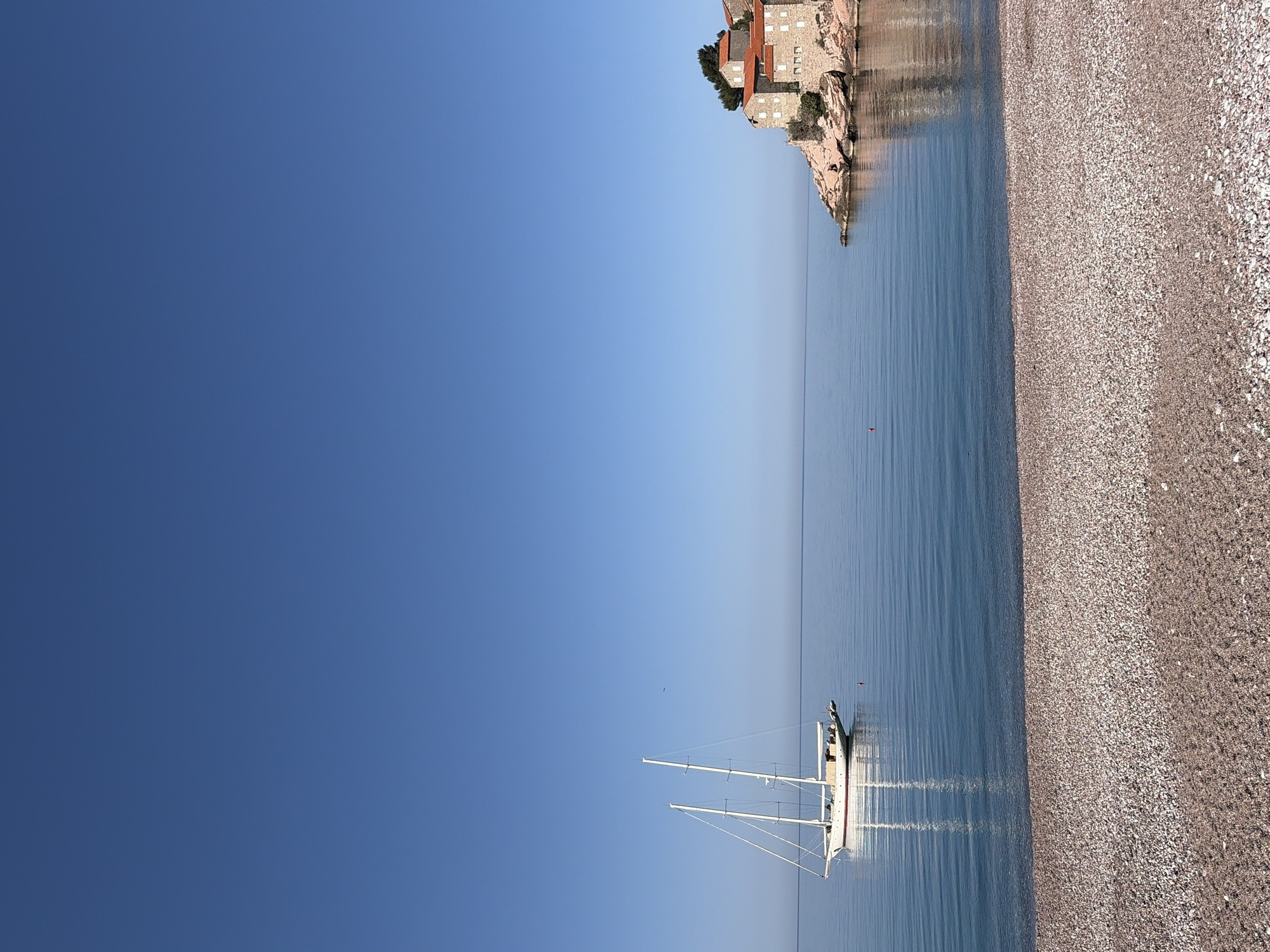 The image displays a serene coastal landscape identified as Sveti Stefan, Montenegro, under a clear, bright blue sky. The picture is oriented vertically, but when viewed horizontally, it shows a calm sea meeting a pebbly beach on the right.
In the upper right portion of the horizontally viewed image, a historic, fortified island settlement, Sveti Stefan, is prominently featured. The buildings are constructed from light-colored stone with distinctive red-tiled roofs, nestled on a rocky outcrop that extends into the sea. Reflections of these structures are visible in the still water.
In the mid-ground and slightly to the left, a white sailboat with two tall masts is anchored in the tranquil, azure water. Its detailed reflection stretches below it on the water's surface, indicating the calmness of the sea. Further out, a small red buoy floats in the water.
The foreground on the right is dominated by a wide, textured beach composed of countless small, light-colored pebbles. The line where the gentle waves meet the shore is distinctly visible.
No specific activity or event is actively taking place; the scene conveys a sense of peaceful quietude. The bright, clear sky and the quality of light suggest it is daytime, likely late morning or early afternoon. The weather appears fair and pleasant. There is no visible text in the image.