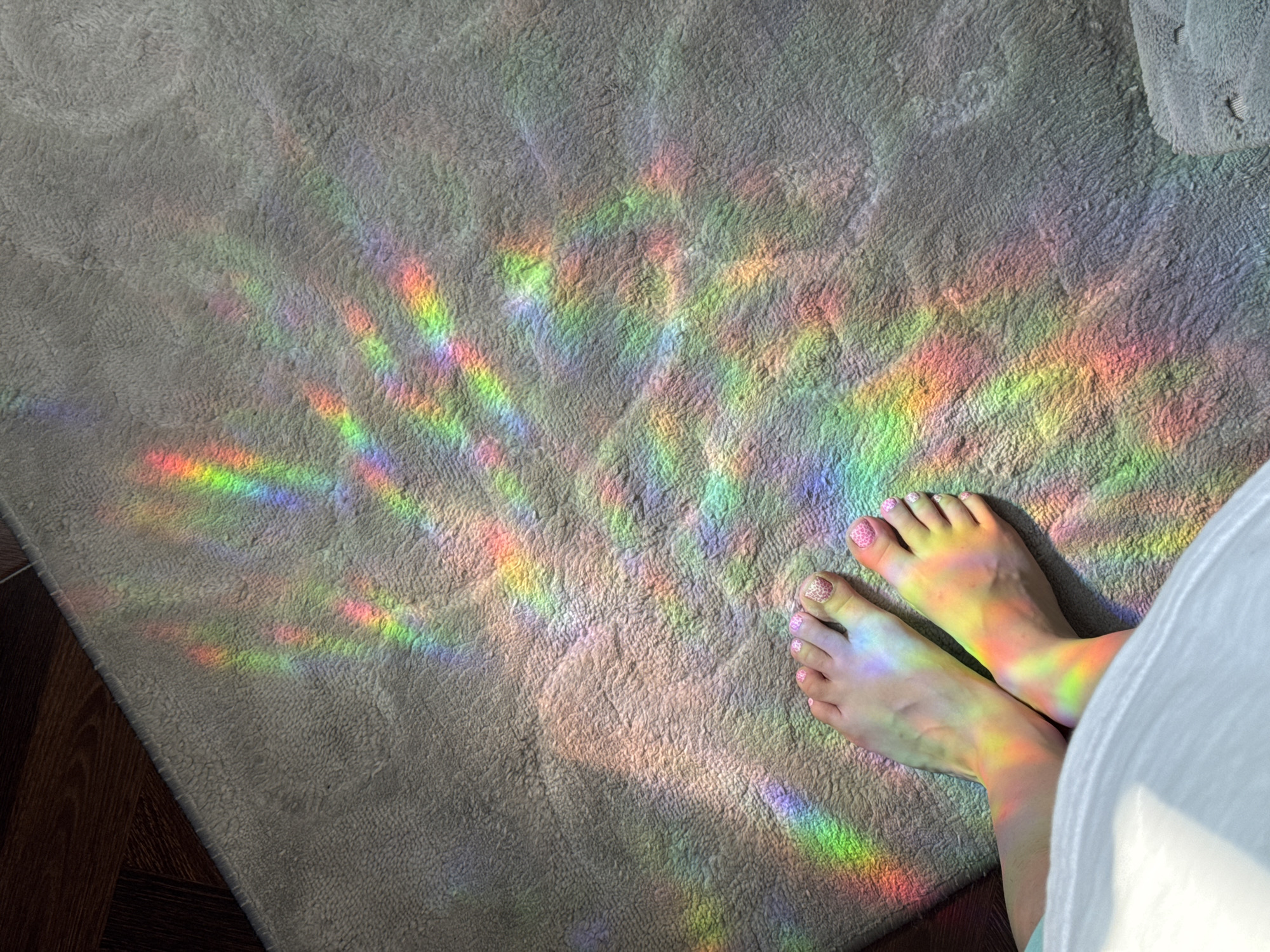 The photograph shows a person's bare feet on a light grey, textured rug. Rainbow light prisms are cast onto the rug and the person's feet. Part of a white garment is visible on the right side of the image. The location is not discernible from the provided image.