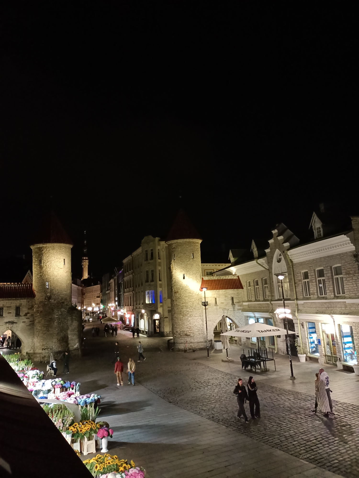 The image depicts a nighttime scene in Tallinn, Estonia, likely in the Old Town, given the medieval architecture. Two prominent, illuminated stone towers with conical roofs dominate the left side of the frame, suggesting they are part of the city's historic fortifications. A cobblestone street stretches out between these towers and rows of historic buildings lining the right side of the image. These buildings are lit from within and from external streetlights, casting a warm glow on the street.
Several people are visible on the street, engaged in various activities. A group of people appear to be walking away from the viewer and towards the illuminated towers. On the right side, a few individuals are walking along the street, and a couple is standing near the entrance of a building. In the foreground, on the left, a flower stall is set up, displaying a vibrant assortment of colorful flowers, including sunflowers and hydrangeas. Some tables and chairs are arranged under a white umbrella in front of one of the buildings on the right, indicating an outdoor seating area for a restaurant or cafe. The overall atmosphere is one of a lively European city at night, with a blend of historic charm and modern activity. The sky is dark and clear, with no visible celestial bodies.