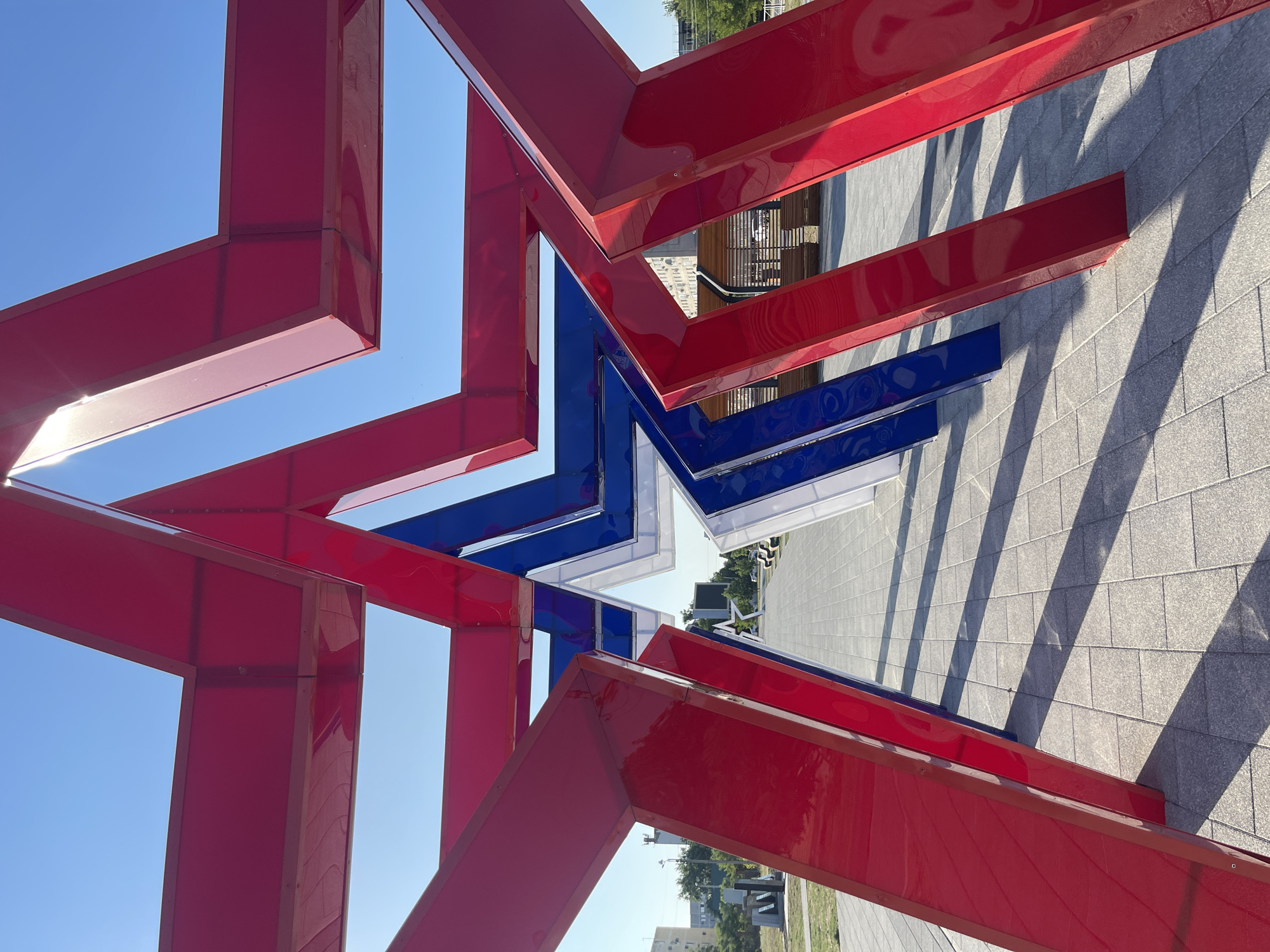 A wide-angle, eye-level shot captures a large, stylized star sculpture constructed from red, white, and blue panels, set against a clear blue sky. The sculpture is the focal point, with its geometric, angular design casting long shadows across a paved walkway composed of square grey tiles. The perspective is from within the sculpture, looking through its layered, star-shaped openings.
In the background, beyond the sculpture, a park-like setting is visible. There are trees, some buildings with windows, and benches under a wooden canopy structure. The overall scene suggests an outdoor public space, likely a park or plaza. The lighting indicates it is daytime, with the sun casting sharp shadows. There are no people visible in the immediate foreground or mid-ground of the image.
The sculpture itself is abstract and modern. The red, white, and blue colors are vibrant. The structure appears to be made of a smooth, reflective material. The tiling on the ground is uniform and well-maintained. The buildings in the background appear to be typical of an urban or suburban environment. The overall impression is one of a public art installation in a well-lit outdoor area.
The location is identified as the city of Budyonnovsk, Russia, specifically at the memorial complex dedicated to the heroes of the Great Patriotic War. The sculpture is part of this memorial complex.
