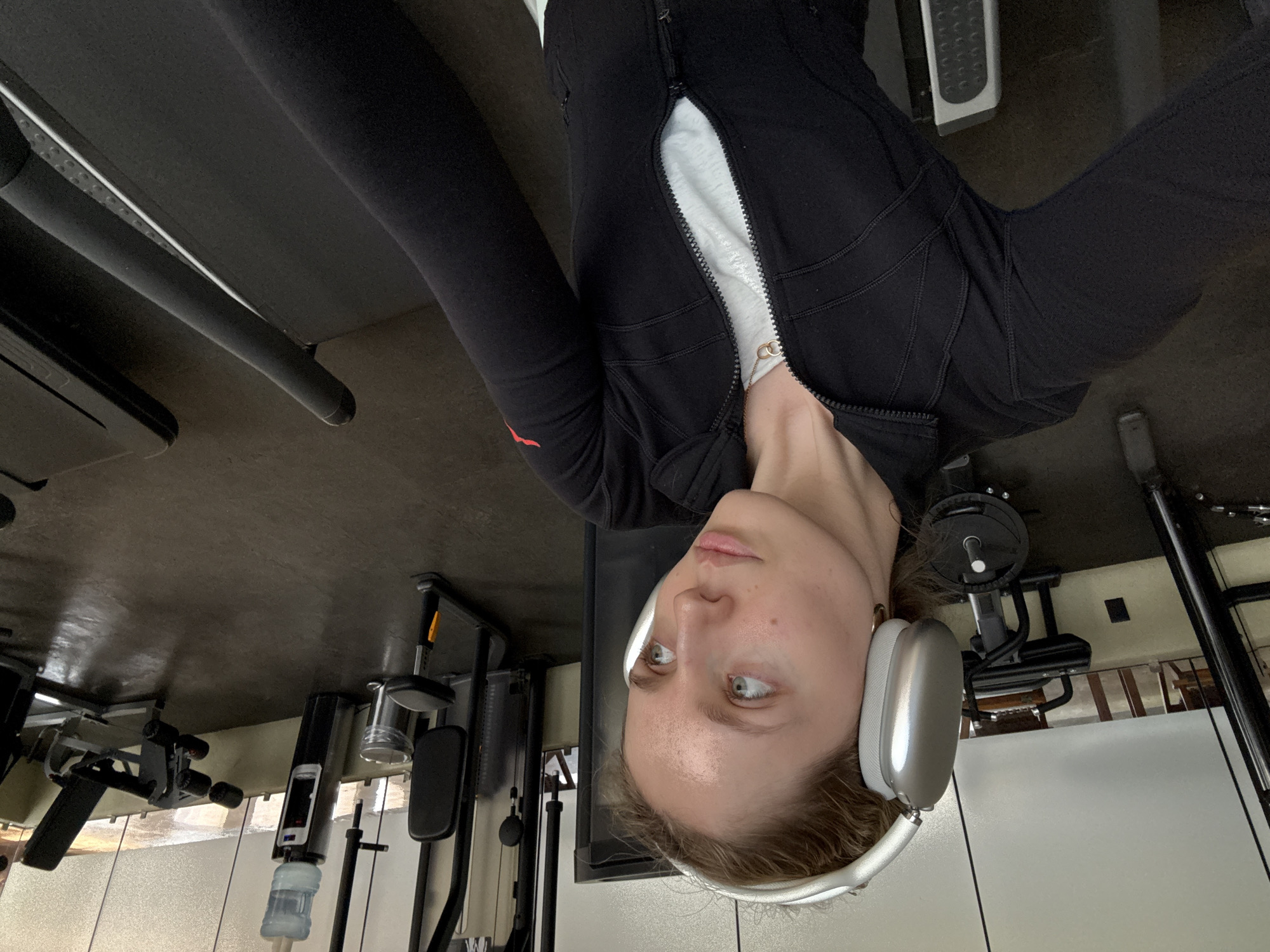A woman is shown wearing headphones, a black jacket, and a white shirt in a gym. The gym appears to be located in Mexico City, Mexico, given the architectural style of the building's walls and the overall aesthetic.