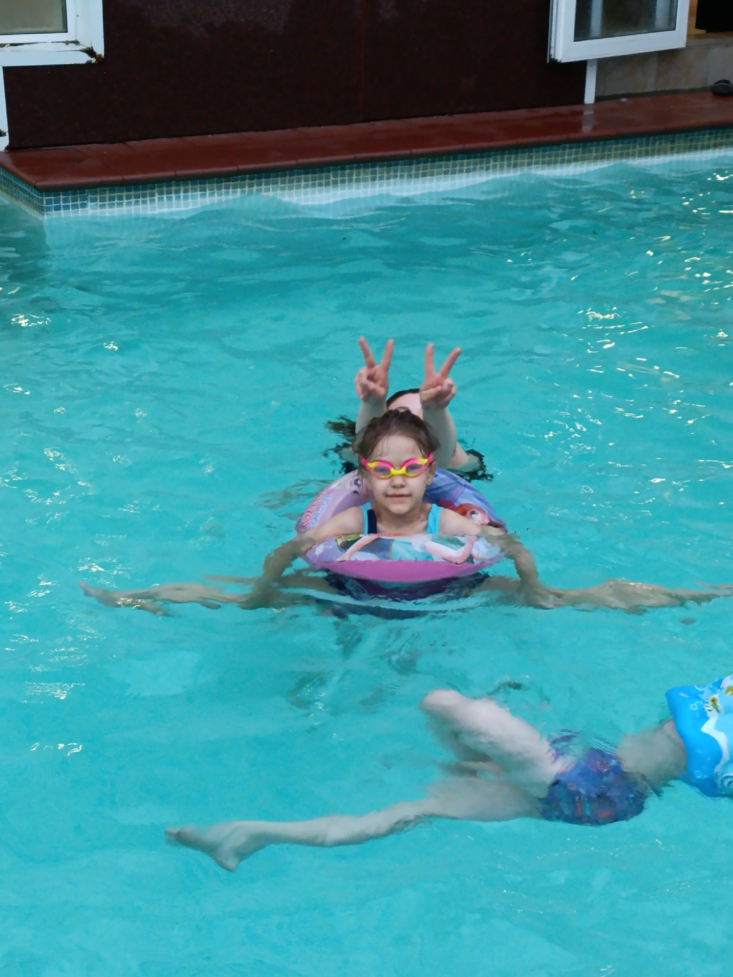 The image depicts an indoor or semi-enclosed swimming pool environment with multiple individuals. No specific visual cues within the image indicate its geographic location in Chaikovsky, Russia, or any other city.
The pool features light blue water with visible ripples. The pool edge consists of small blue and white mosaic tiles at the waterline, topped by dark red coping stones. In the background, a dark brown wall, possibly part of a building, is visible, containing two white-framed windows; one partially visible on the far left and another more complete window centered towards the right.
In the mid-ground, a young girl, estimated to be between 5-8 years old, is positioned upright in the water. She is wearing yellow and pink swimming goggles and a blue swimsuit top. A pink inflatable swim ring, adorned with cartoon characters (likely from Disney's Frozen), supports her around her waist. She looks directly at the camera with a slight smile. Behind her, an adult or older child's head is partially visible above the water, with both hands raised, forming a "V" or peace sign gesture. The girl's legs are extended submerged in the water.
In the lower right foreground, a second young child, likely a toddler or preschooler, is mostly submerged, floating or paddling on their stomach. This child wears blue patterned swim trunks, and a blue inflatable arm float is visible on their right arm. Their legs are also visible beneath the water's surface.