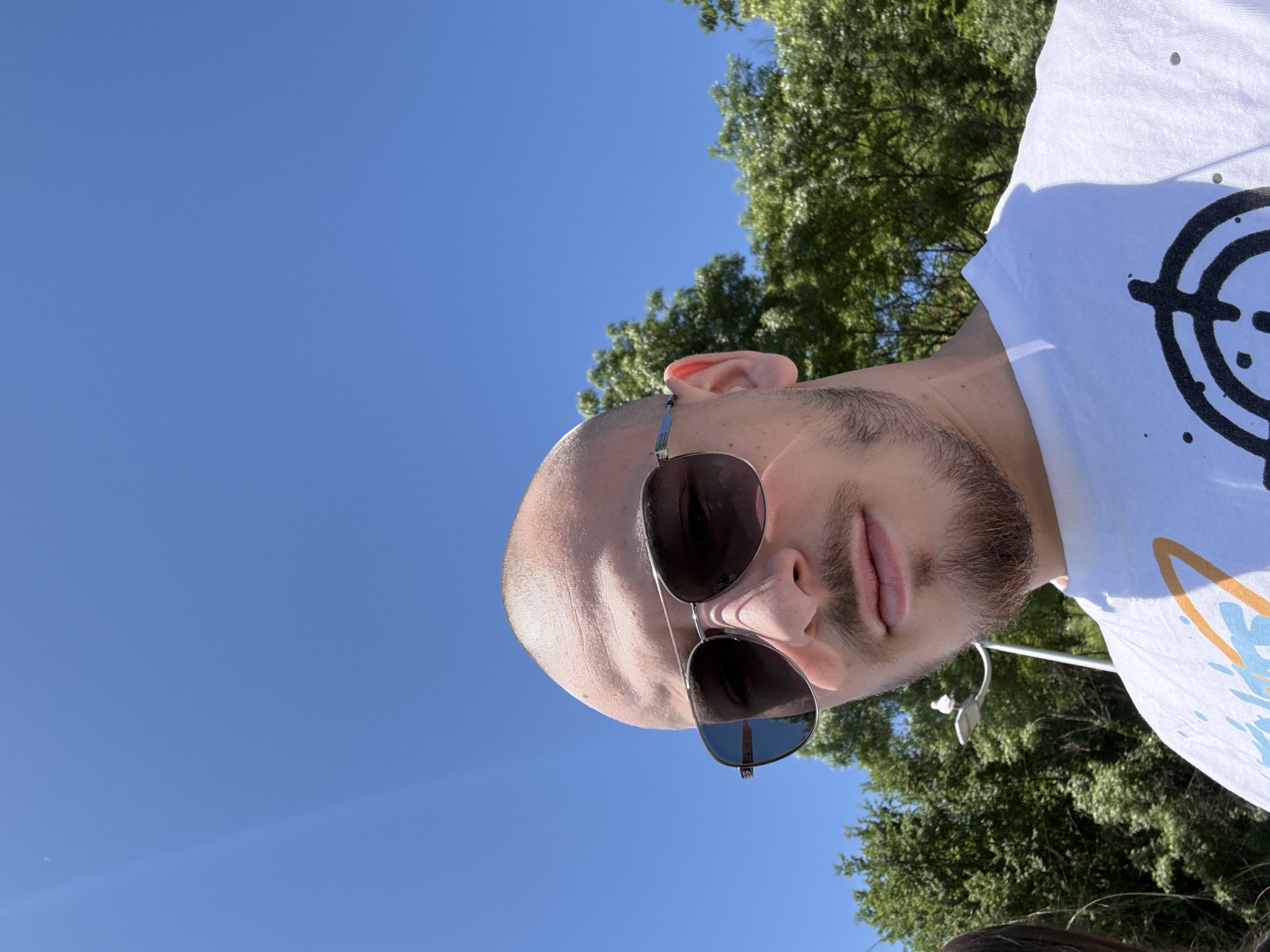 The image is a low-angle selfie of a bald man with a beard wearing sunglasses, taken outdoors on a sunny day in Burgas, Bulgaria. The background includes a clear blue sky and the foliage of trees. The man is wearing a white t-shirt with a graphic design. The photo is oriented vertically and slightly tilted.