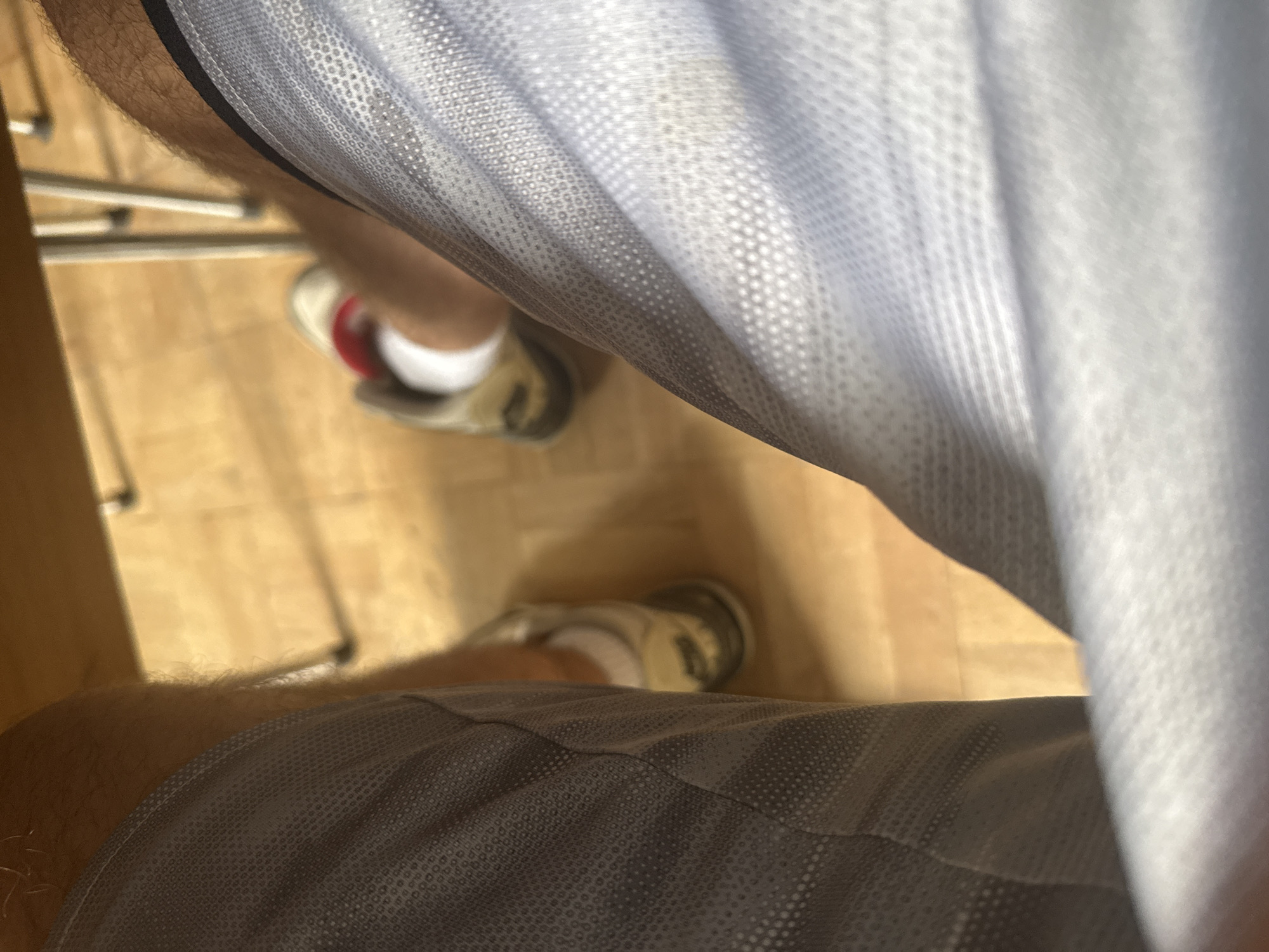 The image shows a person's legs from a first-person perspective. They are wearing grey athletic shorts and white socks with athletic shoes. The shoes appear to be white and gold with red accents. The person is sitting at a wooden table, and the floor is made of light-colored wood planks. The scene is indoors, likely in a casual setting such as a home or a cafe. The location of this scene within the city of Sofia, Bulgaria, cannot be determined from the provided image.