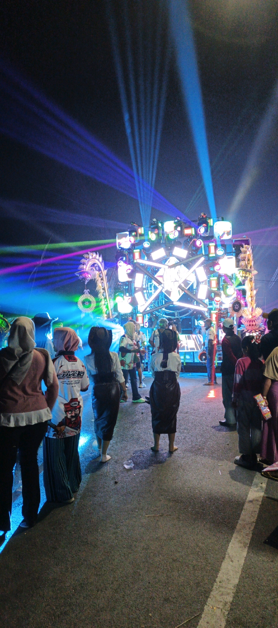 The image depicts a nighttime outdoor event in Kasiyan, Indonesia, characterized by vibrant laser light shows and a large, illuminated stage setup. A crowd of people is gathered, facing the stage. Among the visible individuals are several women wearing headscarves and modest clothing, some with long skirts or dresses. One woman in the foreground is wearing a white shirt with graphics and text. Another woman is wearing a black skirt and white shirt. The environment is an asphalt road, with some debris visible on the ground. The stage is adorned with numerous lights and has a complex, multi-faceted structure. The laser beams cut through the dark sky in various colors, including blue, purple, and green, creating a dynamic and energetic atmosphere. The overall scene suggests a musical performance or a similar entertainment event taking place in the city.