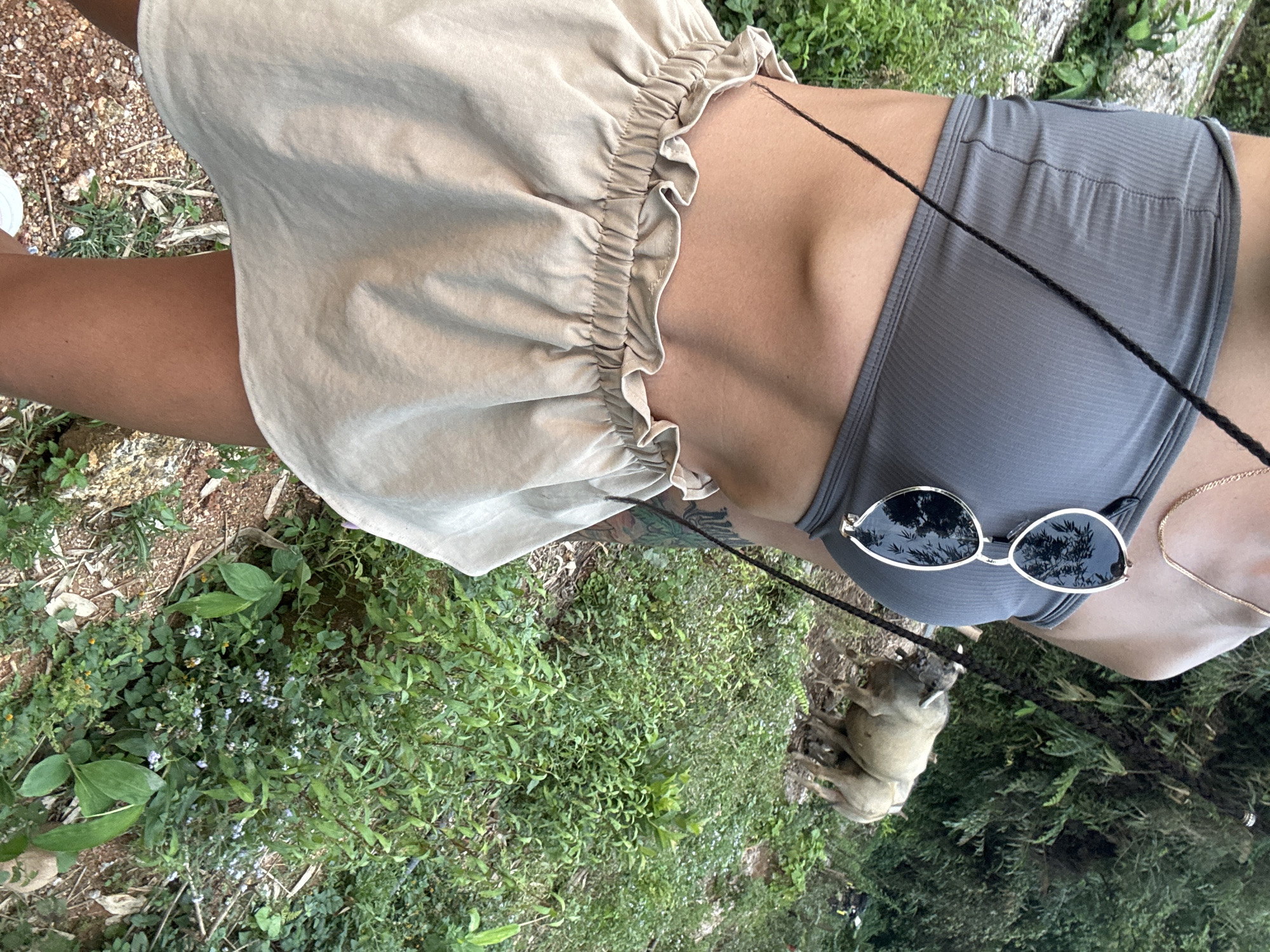 An outdoor, eye-level shot captures a person and their dog in a natural setting within Nậm Dịch Commune, Vietnam. The image focuses on the mid-section of a person, who is wearing a grey ribbed sports bra or crop top and light beige ruffled shorts or a skirt. A pair of stylish gold-framed sunglasses with dark lenses hangs from the neckline of the top, reflecting the surrounding greenery and possibly the sky. A thin, braided black string, likely a dog leash, extends from the person's hand towards a small, fawn-colored dog, possibly a pug, visible in the lower right portion of the frame, partially obscured by foliage. A delicate golden chain is also visible around the person's neck, and a tattoo can be seen on the person's right arm, peeking from under the shorts. The ground is a mix of rich green vegetation, including various leafy plants and small light purple flowers, interspersed with patches of reddish-brown earth and gravel. The bright, natural light suggests it is daytime. No text is visible in the image.