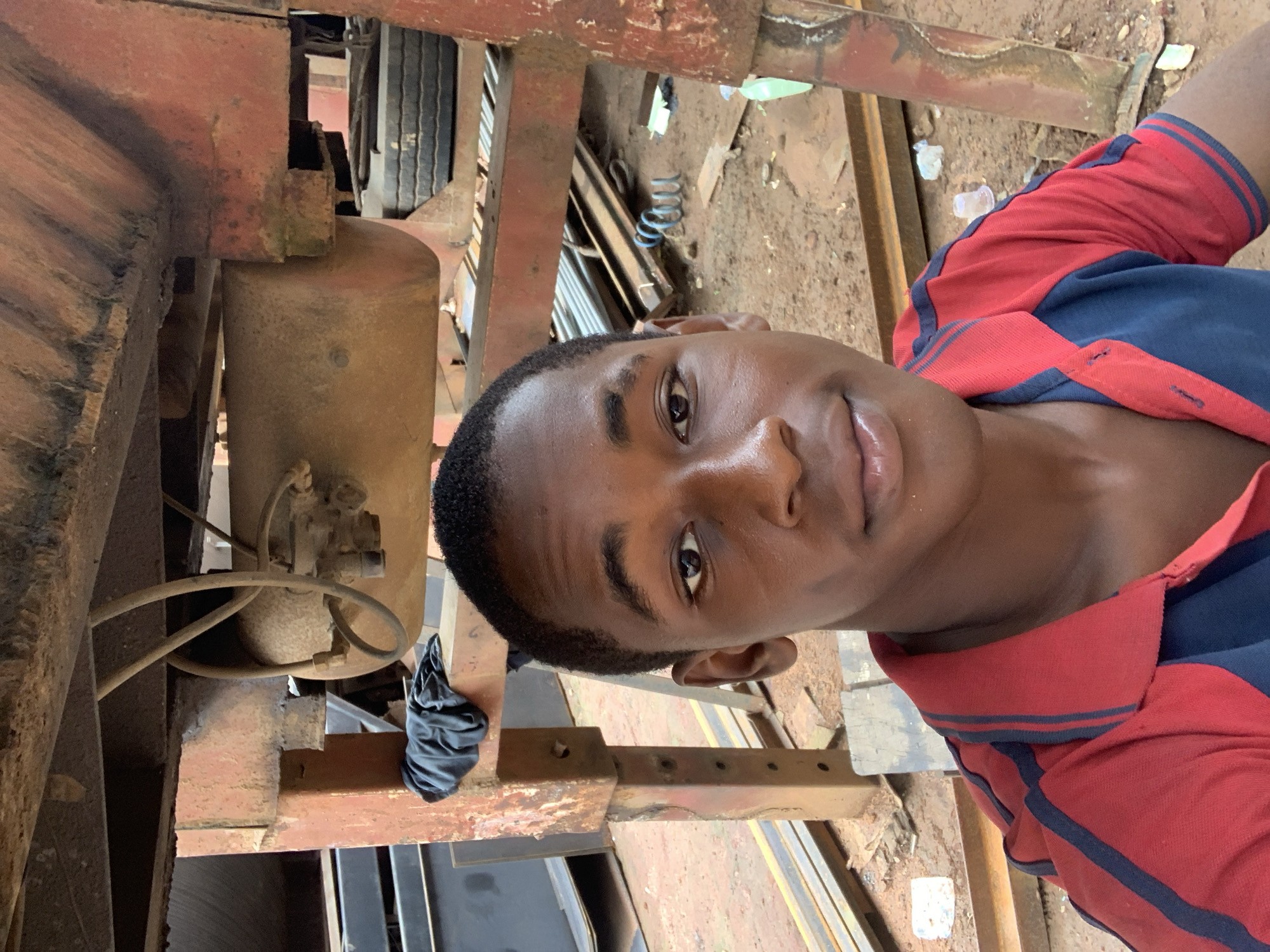 A young Black male, appearing to be in his late teens or early twenties, takes a selfie outdoors. He is positioned in the foreground, his head and upper torso filling the frame. He has short, dark hair, dark brown eyes, and smooth, dark skin. He is wearing a red polo shirt with dark blue accents on the collar and stripes on the sleeves. His expression is neutral, and he is looking directly at the camera.
The background suggests an industrial or workshop setting. Dominant elements include weathered, rusty red metal structures, likely part of a vehicle or machinery. A large, cylindrical tank, possibly a fuel or air tank, is visible suspended from one of the structures, with tubes and wires attached. Black cloth is draped over a metal beam. In the lower portion of the frame, a tire is visible, suggesting it is part of a vehicle. The ground is covered in dirt and scattered with various objects, including metal rods, coiled springs, and some plastic debris like bottles and cups. The overall impression is of a practical, working environment.
While the image is taken in Nkpor, Nigeria, the specific details of the location within Nkpor are not discernible from the visual information provided. The scene appears to be an open-air area within a facility that deals with heavy machinery or vehicle repair/maintenance.