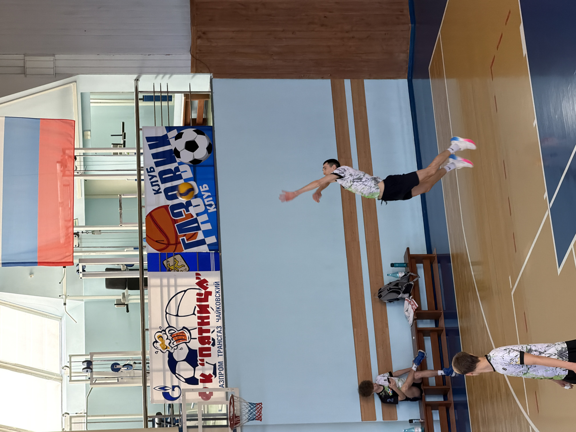 The image depicts the interior of a gymnasium in the city of Chaikovsky, Russia. A young man in a white and black patterned top and black shorts is airborne, performing a gymnastic or acrobatic maneuver. He is suspended above a wooden basketball court. A bench with a backpack and water bottles sits near the wall, where another young man is reclined. A third person, similarly dressed, stands at the lower right of the frame. Banners featuring sports themes (soccer and basketball) and the name "AZPRO PROM TRANSGAZ CHAIKOVSKIY" hang on a wall near an open-air upper level. A Russian flag is also visible. A basketball hoop is attached to the wall.