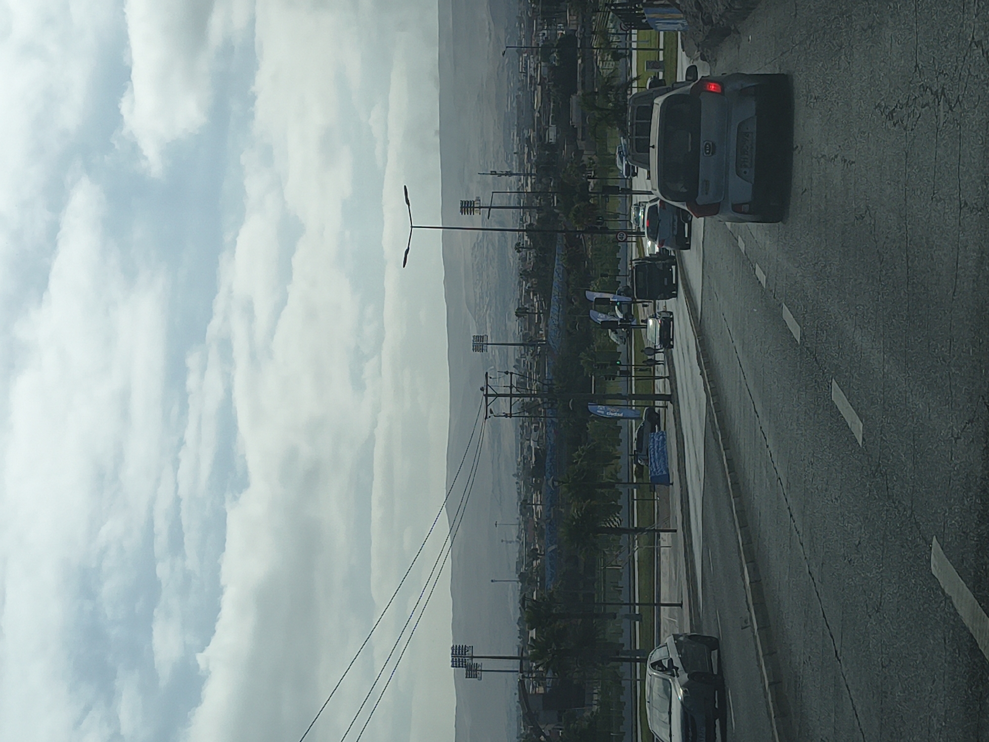 A view from a moving vehicle traveling along a city street in Arica, Chile, on an overcast day. The road, marked with white lines, shows other vehicles in motion. To the side, a park or public space is visible, equipped with lighting fixtures and utility poles, suggesting urban development. In the background, mountains stretch along the horizon.