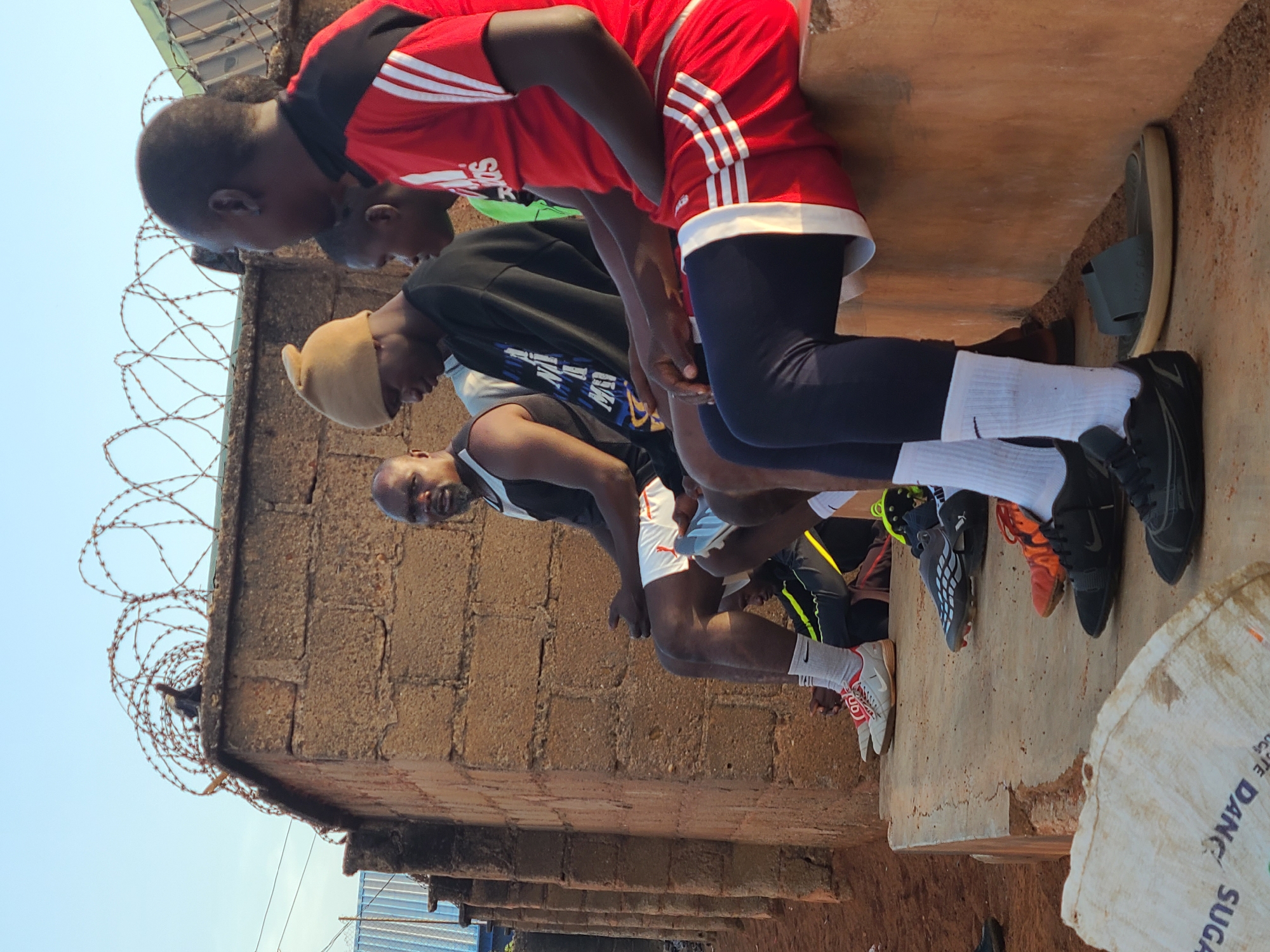 The image captures a group of at least four young men positioned along the edge of a concrete or stone structure. The men are dressed in athletic attire, including jerseys, shorts, leggings, socks, and sneakers. One individual wears a beanie. The setting appears to be outdoors, beside a brick wall topped with barbed wire. A bag labeled "SUGA" is visible in the foreground. The location is in the city of Unknown, Unknown.