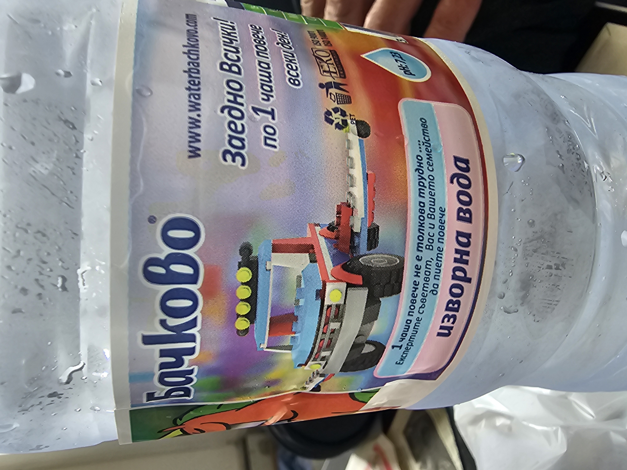 The image displays a clear plastic water bottle covered in condensation, suggesting it is cold. The bottle features a colorful label, predominantly showing the brand name "Бачково" (Bachkovo) in large blue letters. The label's design includes a vibrant illustration of a toy-like truck, possibly made from building blocks, featuring red, blue, and yellow elements.
Visible text on the label is in Bulgarian and includes motivational phrases about hydration: "Заедно Всички! по 1 чаша повече всеки ден!" (Together Everyone! one more glass every day!) and "1 чаша повече не е толкова трудно.... Експертите съветват, Вас и Вашето семейство да пиете повече" (One more glass is not that difficult... Experts advise you and your family to drink more). The product is identified as "изворна вода" (spring water) with a pH value of 7.23. The website "www.waterbachkovo.com" is also printed on the label.
Certifications and logos visible include "AKO SR001", "ISO 9001", "ISO 14001", an "EKO" logo, and a "PET" recycling symbol. In the blurred background, parts of a person's hand are visible, implying the bottle is being held or has recently been handled. The setting appears to be indoors. The provided context indicates the location is Karnobat, Bulgaria. No specific activity or event beyond the presence of the bottle is discernible.