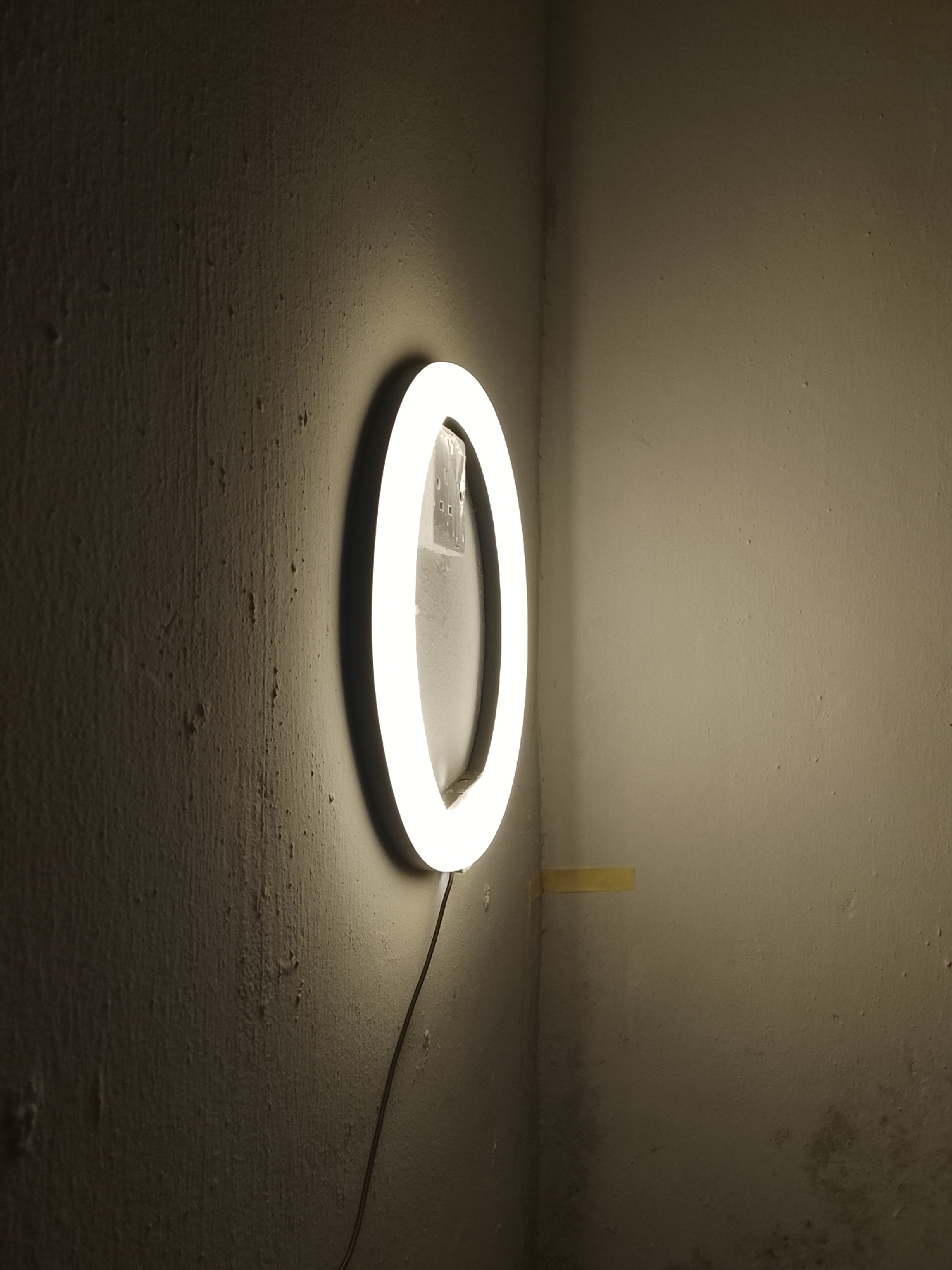 An illuminated ring light in the shape of the numeral zero is mounted on a rough, textured concrete wall in the corner of a dimly lit room. The wall on the left is dark and textured, while the wall on the right is lighter and smoother, with some subtle staining at the bottom. A yellow piece of tape is affixed to the right wall. A black cord extends from the bottom of the ring light, disappearing out of frame. The lighting creates a soft glow around the numeral and casts a shadow on the wall. There are no people or other discernible objects in the scene.