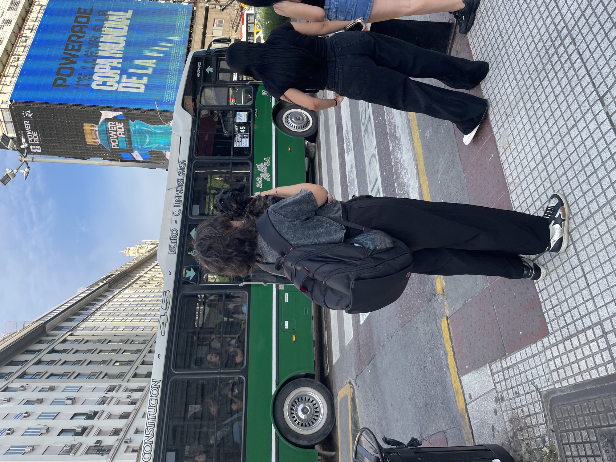 Here is a detailed description of the image:
**Setting:** An urban street scene in Buenos Aires, Argentina. The foreground is dominated by the edge of a street and a crosswalk with people. Vertically, the shot captures the lower portion of a tall building on one side and a towering billboard on the other.
**People:** There are at least three people visible. Two figures are standing near the crosswalk. One has long curly hair, is wearing a grey t-shirt, black pants, and sneakers. The second one is also wearing all black. A third person's lower body is seen; they wear denim shorts and sneakers.
**Objects:**
* **Bus:** A green bus is alongside the street. Text on the side indicates it serves the "RETIRO - C. UNIVERSITARIA" route, and a sign notes the line number. A sticker reads "NUOVOBUS."
* **Billboard:** A large blue billboard has the words "POWERADE TE LLEVA A LA COPA MUNDIAL" in bold lettering. Below, there is a POWERADE ad
* **Building:** A tall building with many windows stands in the background.
* **Street:** The street has a crosswalk with yellow lines.
* **Personal Items:** One person has a black backpack.
**Actions/Interactions:** The people appear to be waiting or standing near the crosswalk. The two people in black are looking toward the bus.
**Environment:** The environment suggests a busy street with public transport and advertisements. The presence of the tall building and large billboard indicates a commercial or central area.