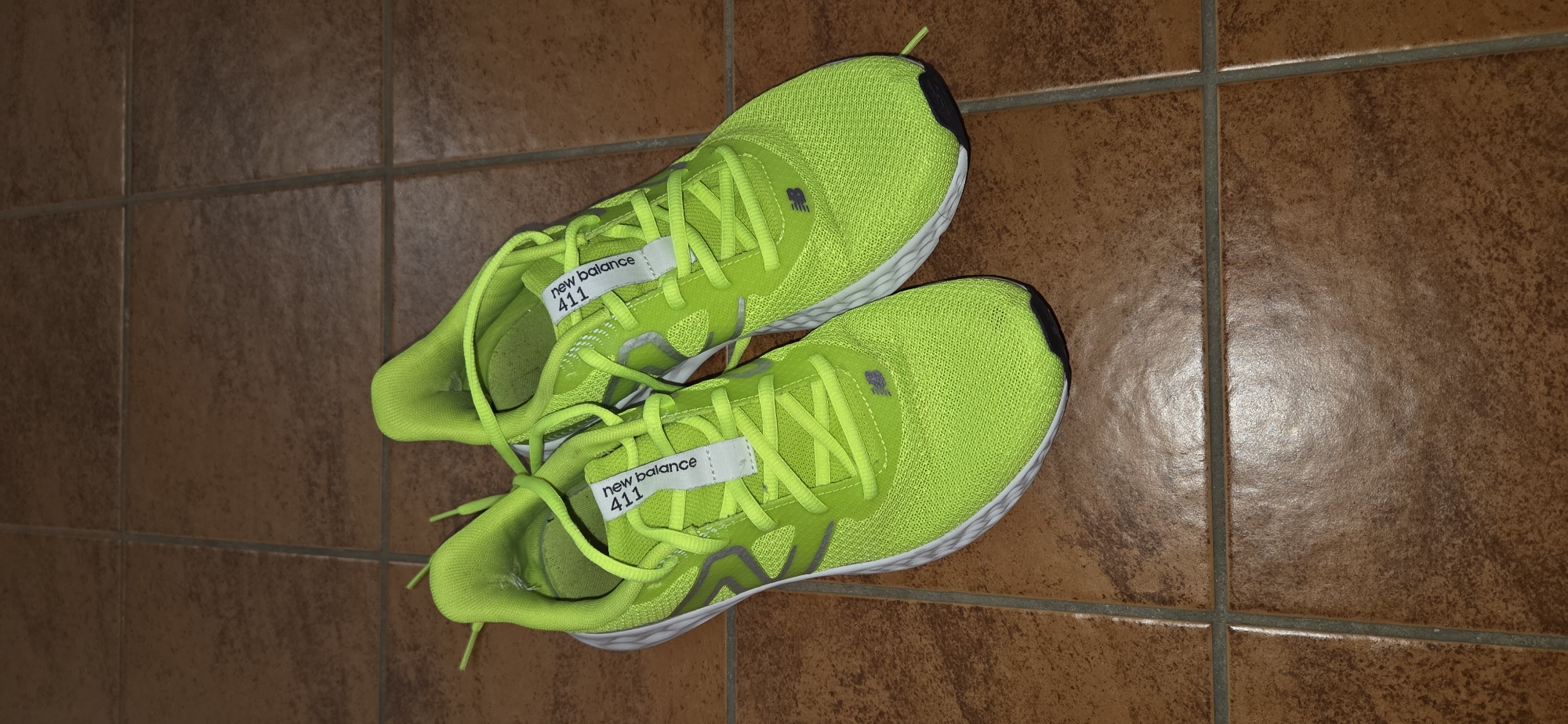 A pair of bright green New Balance 411 running shoes are positioned on a floor tiled with brown, square tiles. The shoes are slightly angled, with the left shoe placed slightly in front of the right. Both shoes are laced with bright green laces. A white label with "new balance 411" is visible on the tongue of each shoe. The lighting suggests an indoor environment, possibly a home. There are no people present in the image. The image does not provide any information about the location in the city of Lipetsk, Russia.