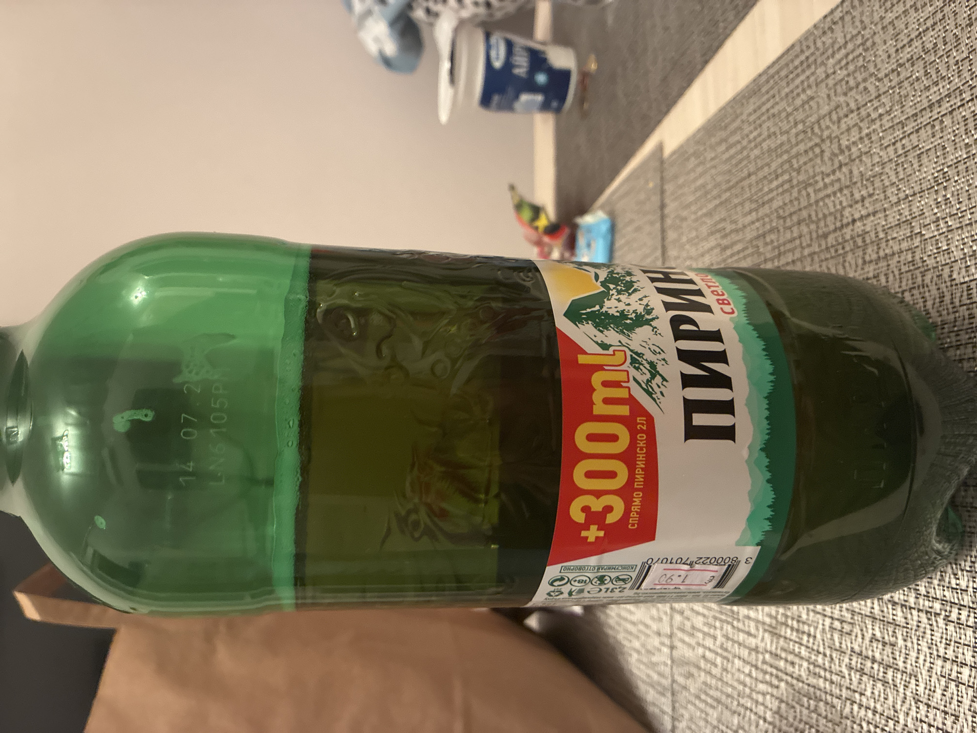 This media file captures a green plastic bottle of beer, lying horizontally on a textured gray surface, likely a table or countertop. The bottle is the primary subject, with its label prominently displayed.
The label identifies the brand as "ПИРИН" (PIRIN), a Bulgarian beer, and specifies "светло" (svetlo), indicating a light beer. A red section on the label highlights a promotional offer of "+300ml" compared to a standard 2L bottle, with "СПРЯМО ПИРИНСКО 2Л" (compared to Pirinsko 2L) written underneath. The total volume is indicated as "2.3LE" (likely 2.3 liters). Responsible consumption symbols are visible, including an "18+" age restriction and the phrase "КОНСУМИРАЙ ОТГОВОРНО" (Consume responsibly).
A price tag on the label shows "€ 1.90" and a barcode with the numbers "38000227010/0". Embossed into the bottle's plastic near the neck, the date "14.07.26" and a code "LN6 105P" are visible. In the blurred background, another container, possibly a dairy product, is faintly visible with the text "АЙРУ" (AIRU). The scene is illuminated by indoor lighting, suggesting a typical home setting in Blagoevgrad, Bulgaria. No people or active events are present.