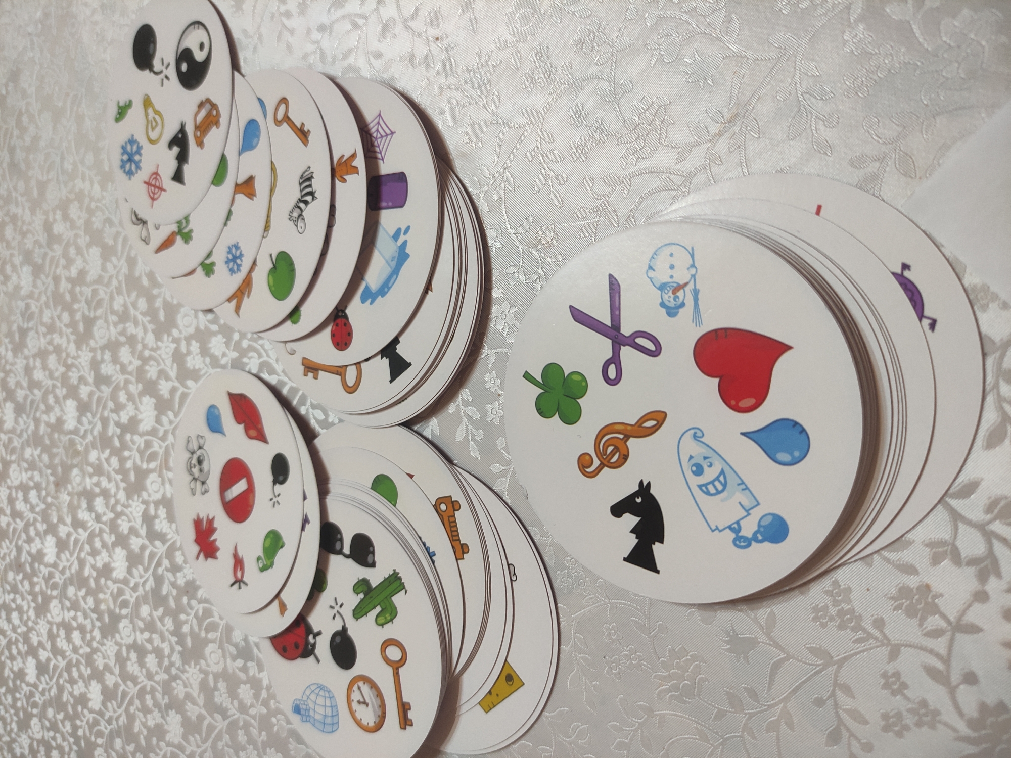 The image shows three stacks of Dobble/Spot It! game cards arranged on a white patterned tablecloth. The top card of the leftmost stack has a yin yang symbol, a ghost, a light bulb, and a small car. The top card of the middle stack has a stop sign, a skull, a leaf, and a flame. The top card of the rightmost stack has scissors, a clover, a musical note, and a black chess knight. The tablecloth has a floral pattern. The location of the image is unknown.