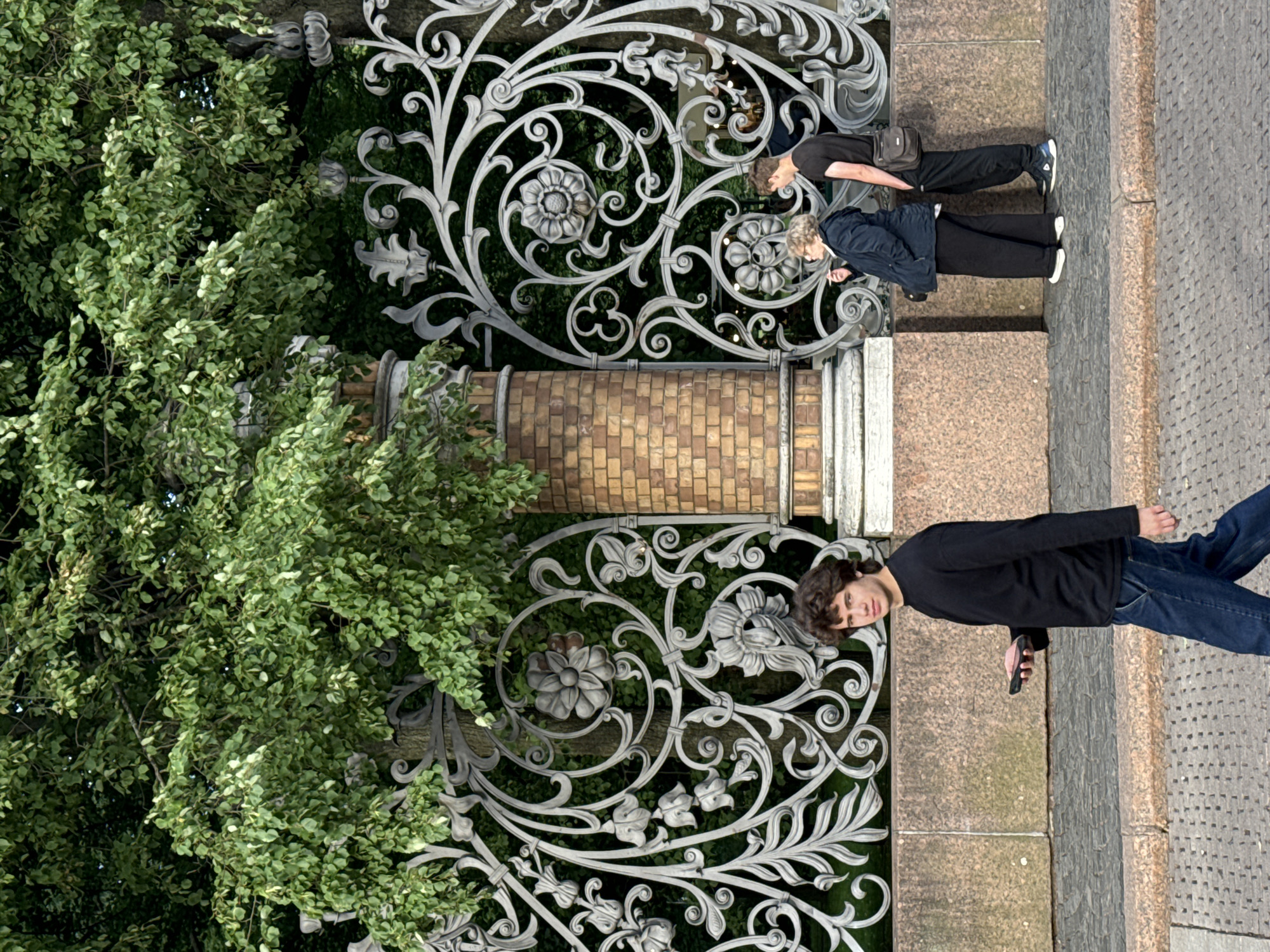 The image shows three people standing near an ornate metal fence and a brick pillar. The fence has intricate scrolling patterns and floral motifs. To the left of the fence is a lush green tree with dense foliage. Behind the fence, dark green bushes are visible.
Two individuals are standing close to the fence, looking downwards. One, wearing a dark jacket and black pants, has short, curly brown hair and appears to be a young person. They are holding a dark object, possibly a phone, in their right hand. The other person, whose face is partially obscured, seems to be wearing glasses and dark clothing. Further back, a third person in a black shirt and dark pants is also visible, with a small dark bag slung across their shoulder.
The ground appears to be paved with stone. The lighting suggests it is daytime, possibly late morning or afternoon, with overcast skies. The architectural style of the fence and the presence of greenery suggest an urban park or garden setting in a city. The context provided indicates the location is Saint Petersburg, Russia. No visible text is present in the image.