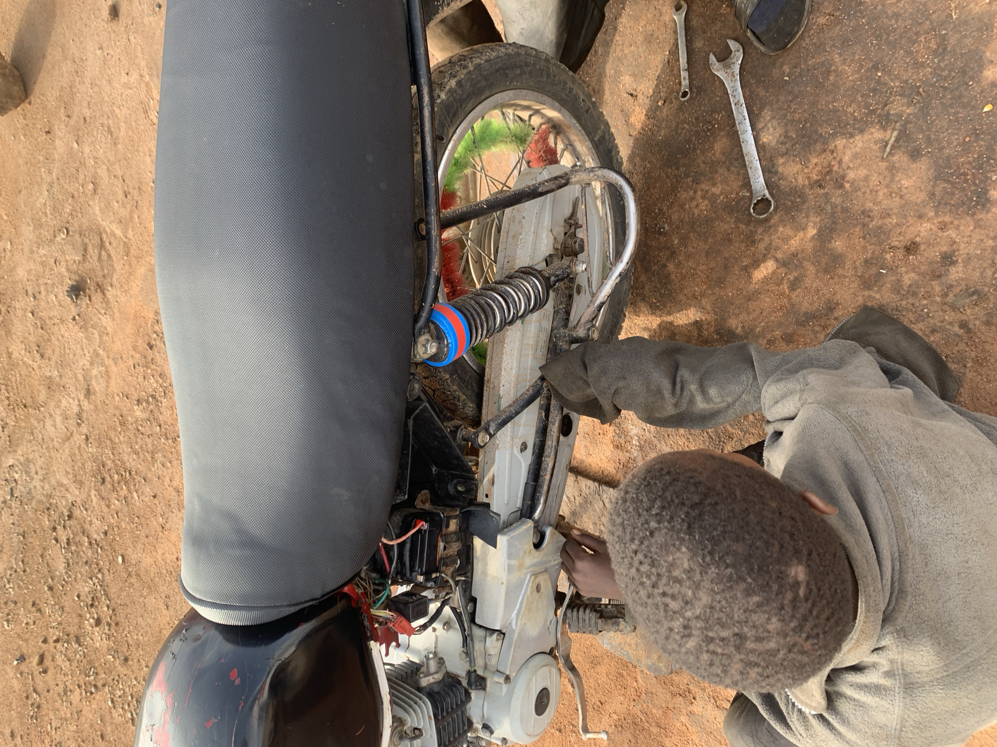 This overhead image captures a person engaged in motorcycle repair in an outdoor setting in Barakin Lerawi, Nigeria. The scene is characterized by dry, reddish-brown dirt ground, typical of a dusty, possibly rural or semi-urban environment.
The primary subject is a young person, seen from above and behind, with short, dark curly hair. They are wearing a gray, long-sleeved top and are leaning over, concentrating on the motorcycle's engine. Their right hand is actively working on components within the exposed engine area.
The motorcycle itself appears to be a small-displacement scooter or underbone model. Key visible parts include a black, textured seat that dominates the left side of the frame, and a partially visible red and black painted bodywork with some peeling paint. The engine block, featuring white or silver cooling fins, is exposed, along with a tangle of wires and electrical connections. A rear wheel with spokes and a tire is visible behind the engine, as well as a rear shock absorber with a black spring and distinctive blue and red plastic components. A silver-colored frame element or exhaust pipe is also present near the rear wheel. A kickstand can be seen supporting the motorcycle.
On the ground, two open-ended wrenches of varying sizes are laid out, indicating ongoing mechanical work. A dark sandal or flip-flop is also partially visible near the tools. The bright, natural lighting suggests the activity is taking place during daytime under clear weather conditions. No text is visible in the image. The overall scene depicts practical, hands-on motorcycle maintenance.