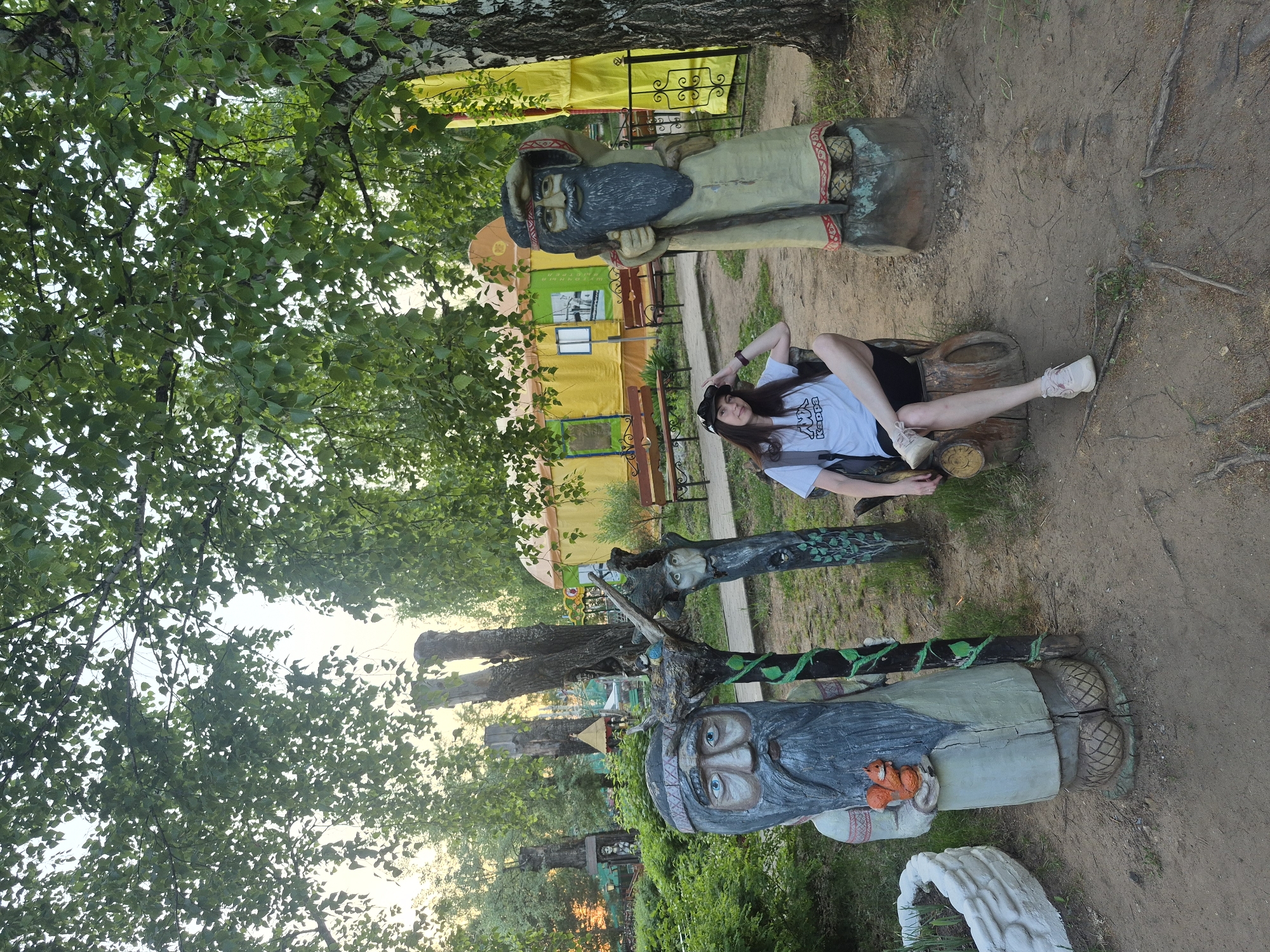 A medium shot captures a woman lying on a log next to a wooden statue of a bearded man in an outdoor setting. The statue is positioned to the left of the woman and appears to be a part of a collection of similar wooden carvings. The woman wears a white t-shirt, black shorts, a black hat, and light-colored sneakers. She is lying on her back with one arm behind her head. The background includes trees, other wooden statues, a yellow structure, and dirt ground. The location is in the city of Unknown, Unknown.