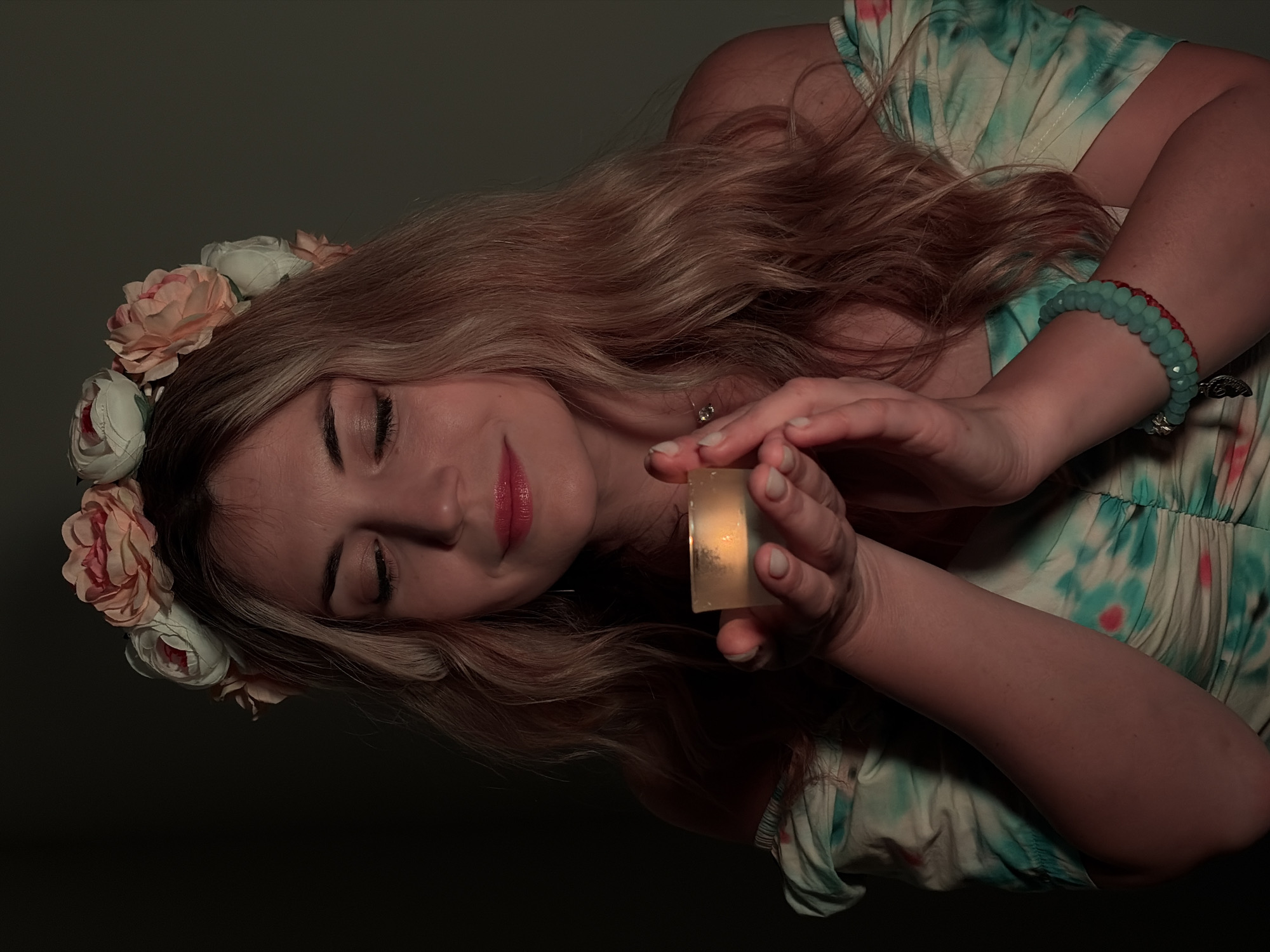 A woman with long blonde hair and a floral crown is holding a lit candle in her hands. She is wearing a tie-dye dress and a bracelet. The background is dark. There is no discernible evidence suggesting a location in Budva, Montenegro.