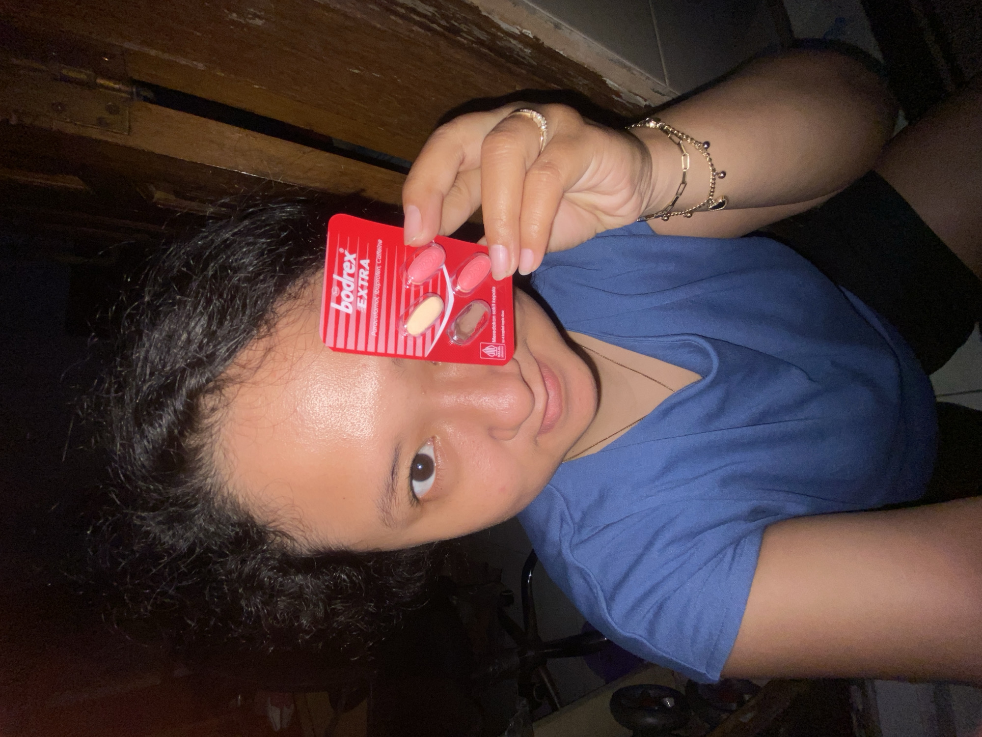 The rotated image captures a person, likely female, looking directly at the camera with a neutral to slightly smiling expression. She has dark hair and dark eyes, and is wearing a blue short-sleeved top and dark shorts. On her right hand, she wears a ring on her ring finger and a gold-colored bracelet on her wrist.
She holds up a red blister pack of medication in front of her face, partially obscuring her forehead. The brand name "bodrex EXTRA" is prominently displayed in white text on a red background with white vertical stripes. Below this, the active ingredients "Paracetamol, Ibuprofen, Caffeine" are listed. Further text in Indonesian, "Meredakan sakit kepala" (Relieves headache), is also visible. The blister pack contains four pills: two are pinkish-red, one is light yellow or cream-colored, and one slot appears empty or is obscured.
The setting is indoors, with a dim background featuring dark wooden structures, possibly furniture or wall elements, consistent with a room. The lighting is low, likely artificial, illuminating the subject's face and the medication. The scene suggests an evening or nighttime moment. The visible text on the medication indicates the context of Gelam, Indonesia.