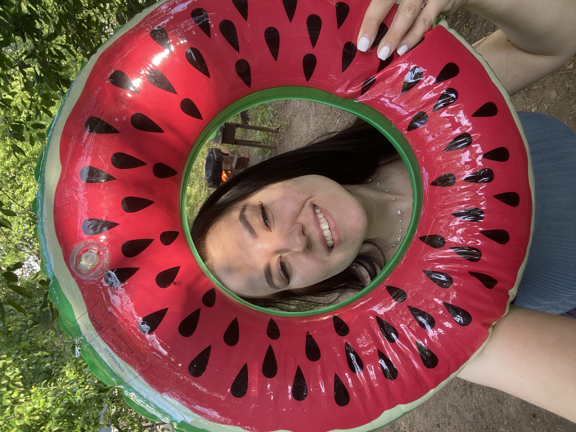 A woman is seen smiling through the hole of a watermelon-shaped inflatable float. The float is red with black seed shapes and a green border. The woman has dark hair and is wearing a necklace. Behind her, visible through the float's hole, is a campfire and some wooden furniture. The surroundings appear to be outdoors, possibly in a park or wooded area, suggesting a recreational setting. There is no indication in the image that this photo was taken in the city of Chaikovsky, Russia.
