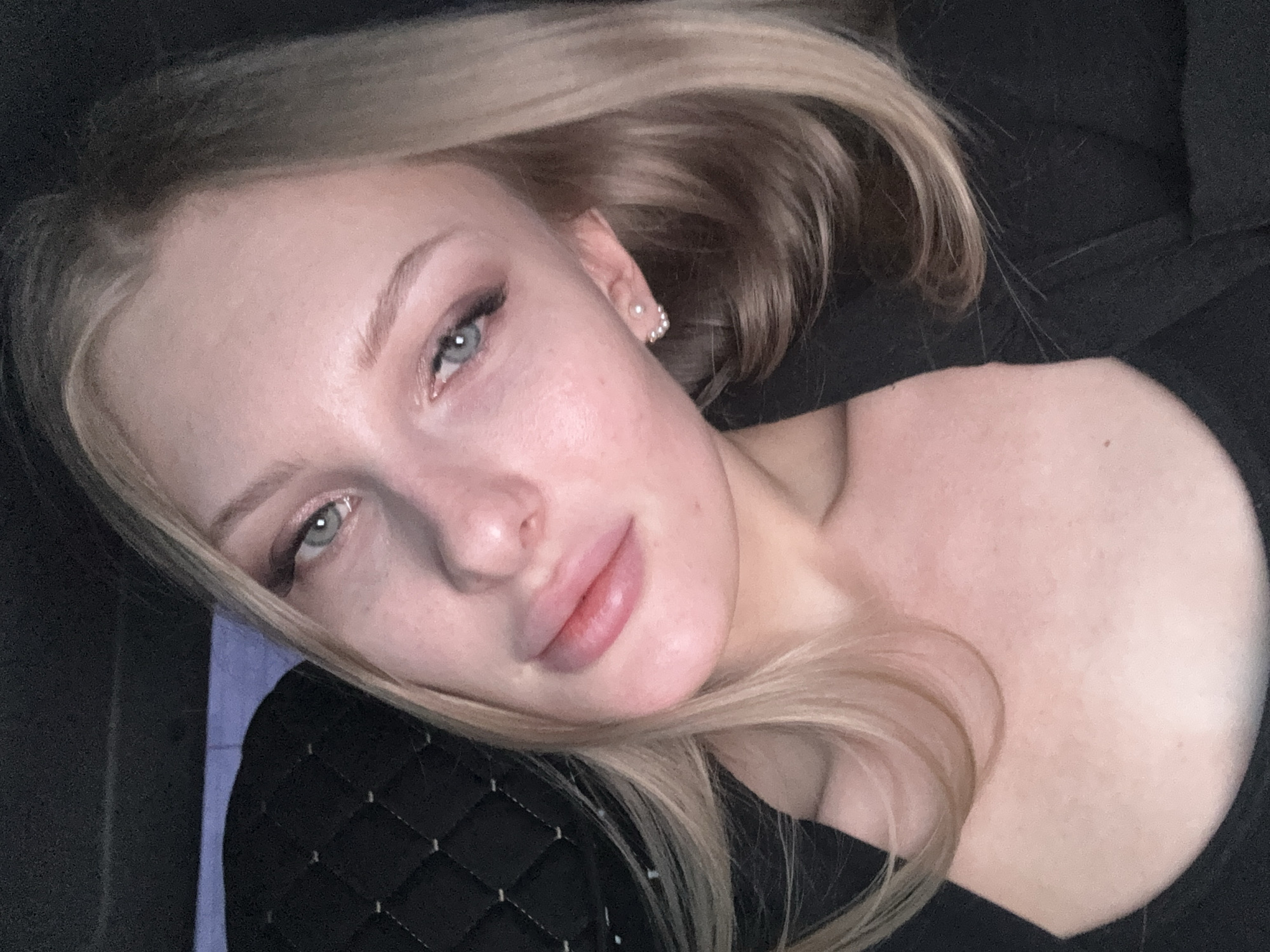 A young, fair-skinned woman with blonde hair is lying down and facing the camera. She is wearing small, pearl-like earrings. She has light makeup on, emphasizing her eyes. She is wearing a black top that appears to be quilted on the left side. The background is dark and blurry, suggesting an indoor setting. There is no indication of location in Perm, Russia.