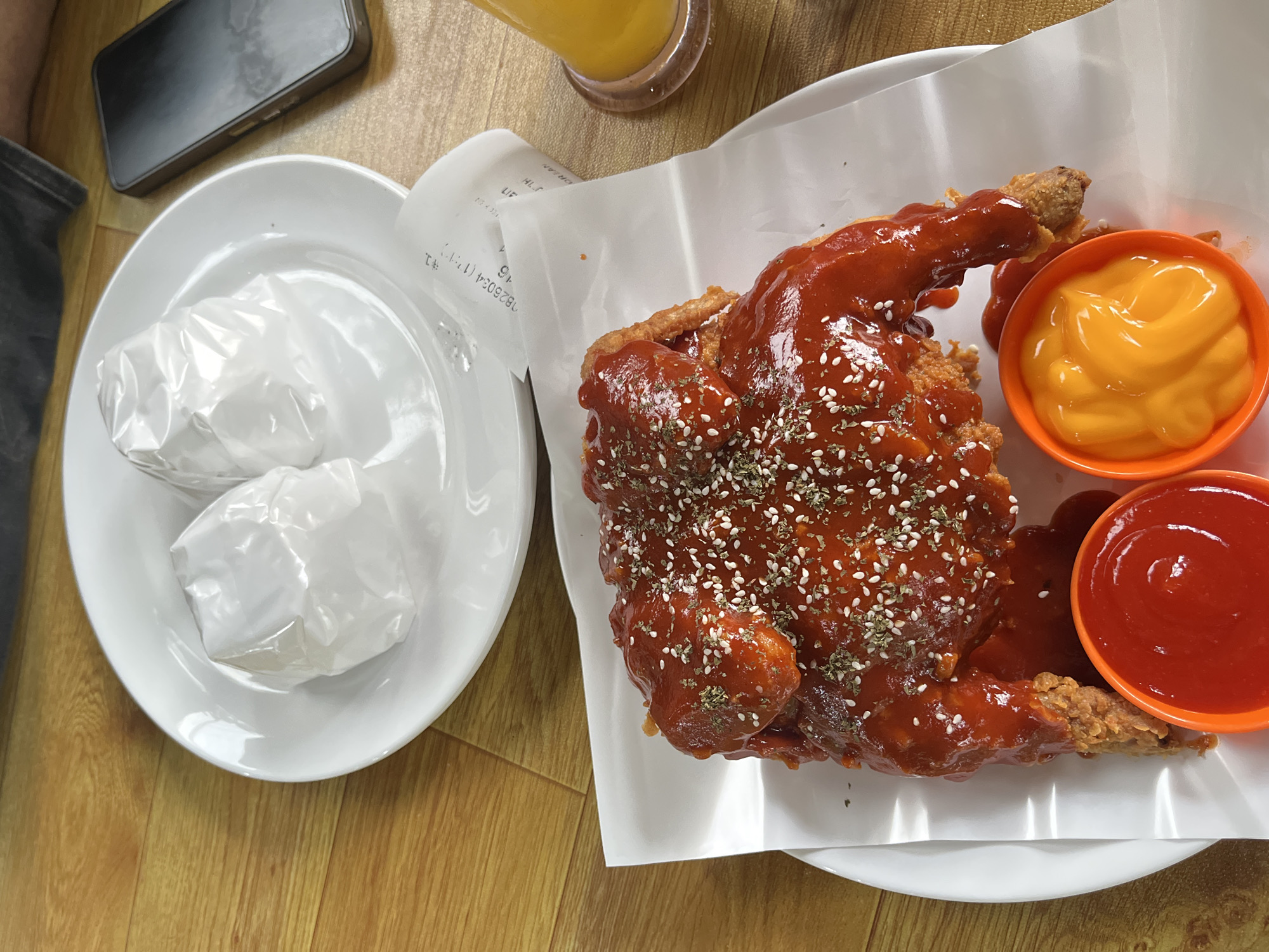A close-up, overhead shot showcases a plate of spicy Korean fried chicken coated in a vibrant red sauce, garnished with sesame seeds and dried herbs. Two small orange dipping bowls, one filled with red sauce (likely ketchup) and the other with a yellow, creamy sauce (possibly cheese sauce), sit in front of the chicken. The chicken is served on a white paper lining atop a white plate, which is placed on a wooden table. To the upper left, another white plate holds two white-wrapped food items, possibly burgers or sandwiches. A crumpled receipt is visible near the plate with the chicken. To the right, a glass of orange liquid, likely juice, rests on a coaster. A smartphone lies on the table to the right of the upper plate. The scene appears to be indoors, with warm lighting. The location specified, Keudeteupinpunti, Indonesia, cannot be visually confirmed from the image alone.
