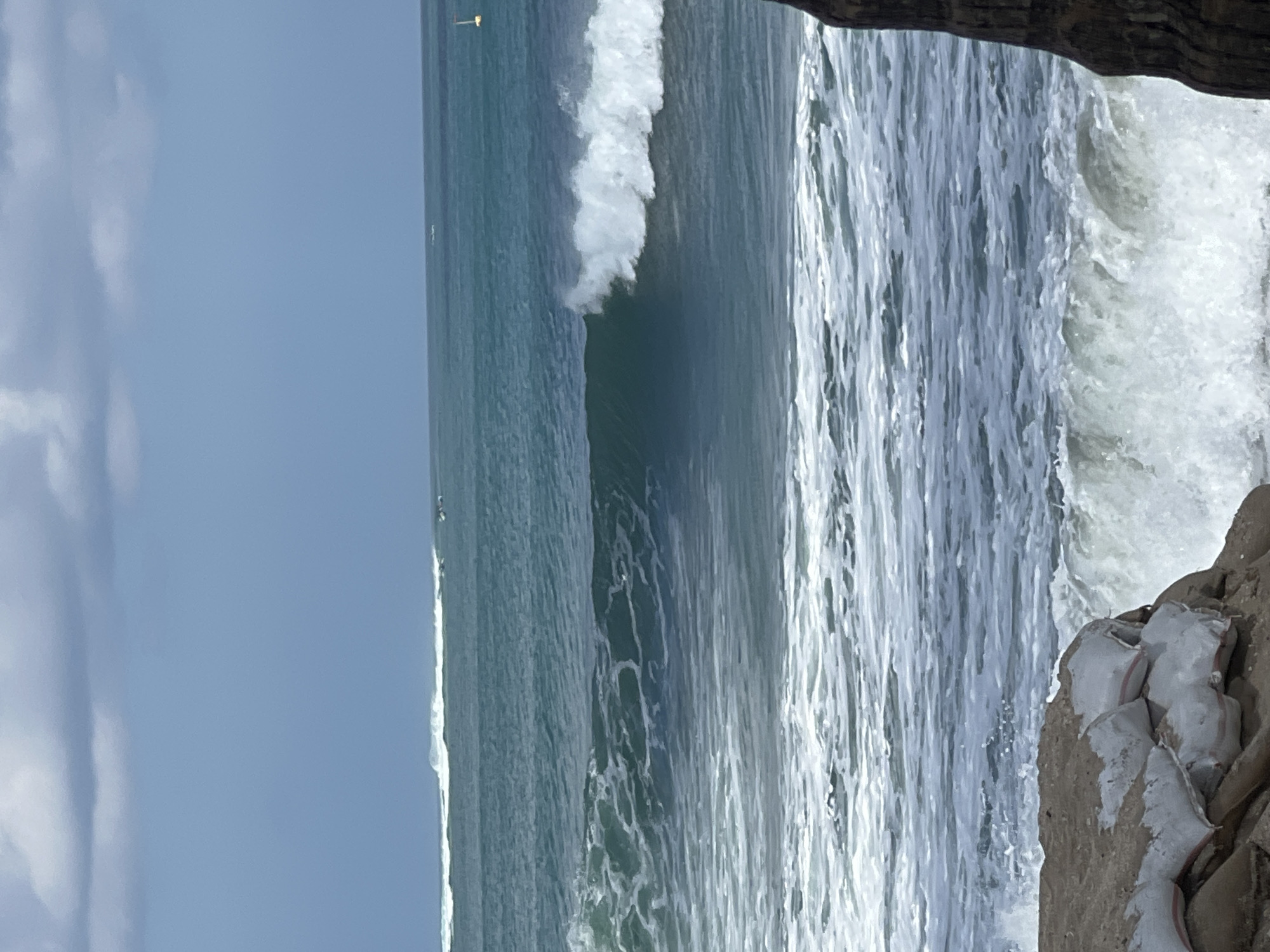 The image depicts a turbulent ocean scene with a breaking wave in the mid-ground. In the foreground, sandbags are stacked on a rocky shore, suggesting coastal defense measures. The water is a deep teal, with white foam from the waves creating a frothy texture. The sky above is partly cloudy, with patches of blue visible. The distant horizon shows a thin line of white surf and what appears to be a small boat. The overall impression is one of a dynamic and potentially rough sea. The image is framed by a dark, textured object on the right side, likely the trunk of a tree. The location is identified as Kuta, Indonesia, a popular tourist destination known for its beaches and surf.
