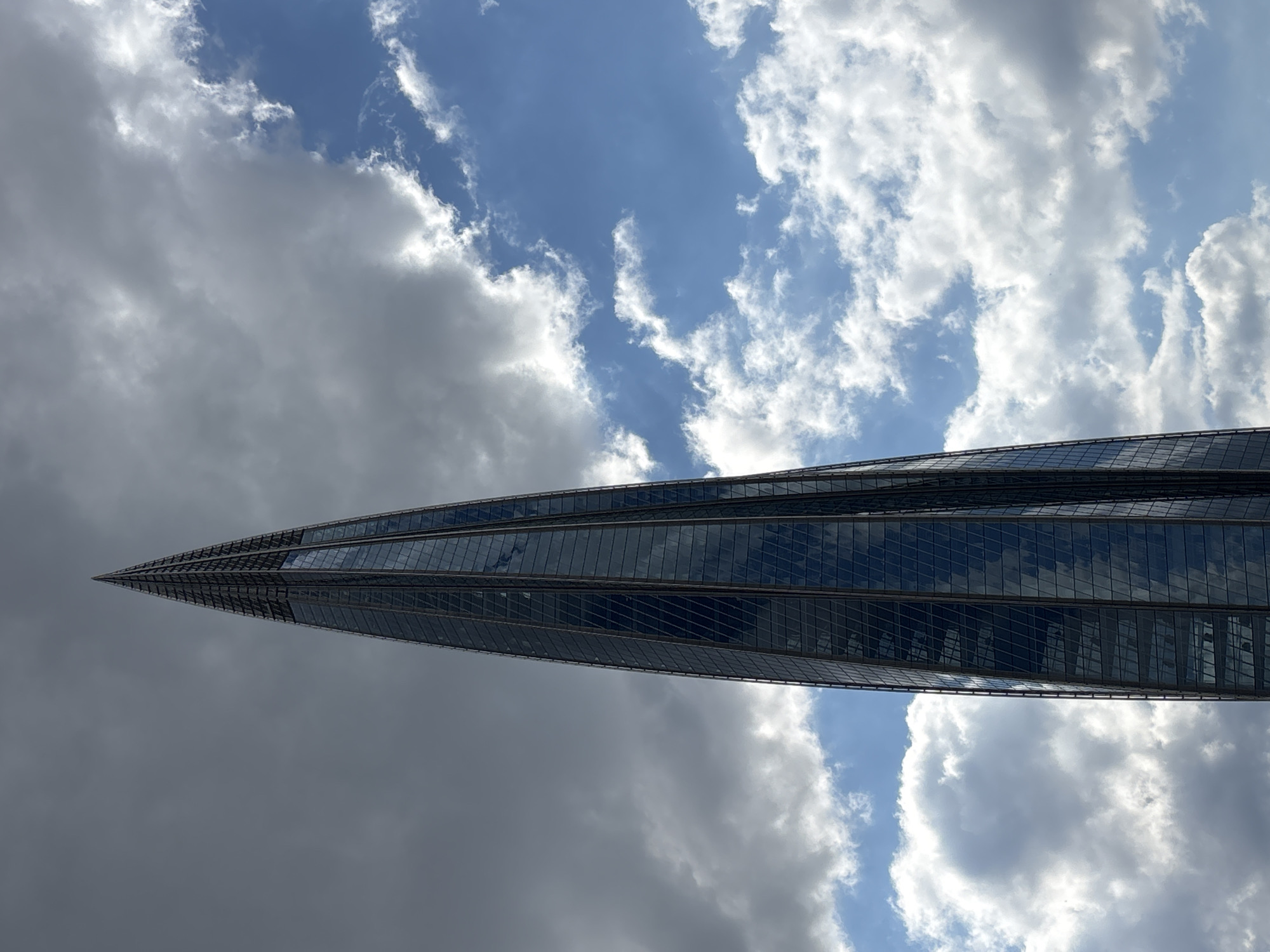 A tall, slender skyscraper with a pointed, spire-like top dominates the frame, reaching towards a cloudy sky. The building's facade is a sleek, dark glass that reflects the surrounding environment. The sky is a dramatic mix of dark, heavy clouds on the left and bright blue sky with white, fluffy clouds on the right. The skyscraper appears to be located in a modern urban setting, given its architectural style. Based on the building's design, it is likely the Lakhta Center in Saint Petersburg, Russia, which is the tallest building in Europe and the northernmost skyscraper in the world. There are no people or animals visible in the image. The image is taken from a low angle, emphasizing the height and imposing presence of the skyscraper.