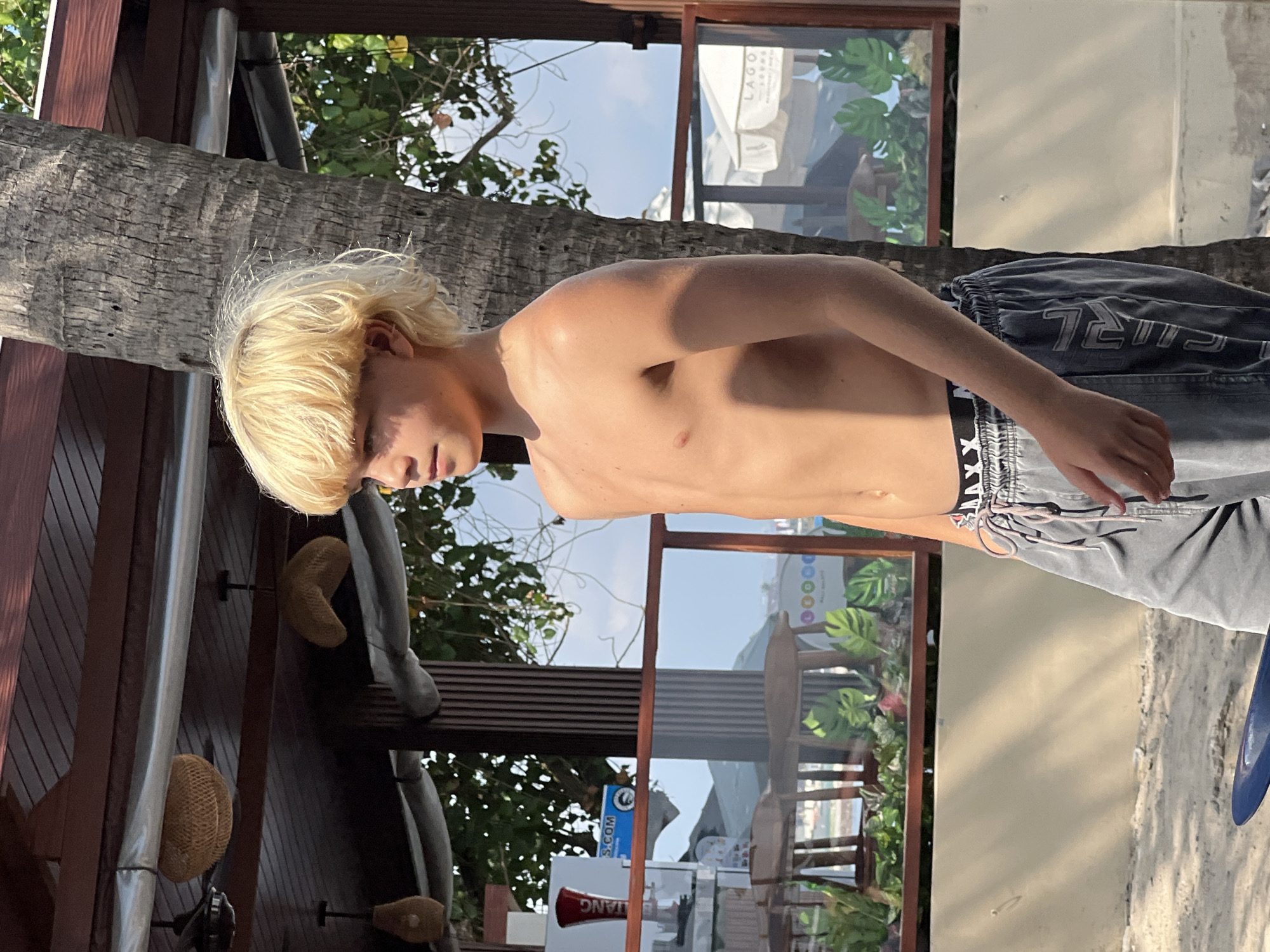 This media file shows a young person with blonde hair, shirtless, wearing dark grey shorts with a black and white patterned waistband. They are standing outdoors, in what appears to be a tropical or beachside setting. The background includes a palm tree trunk on the left and a building with large windows and wooden framing on the right. Through the windows, greenery and what looks like outdoor seating are visible. The sun is casting shadows, suggesting it is either morning or afternoon. The overall mood appears casual and relaxed. Visible text includes "LAGO" on an umbrella in the background and "S.COM" on a blue and white sign. The presence of palm trees and a beach-like environment, combined with the text "Kuta, Indonesia," suggests the location is likely Kuta, Indonesia.