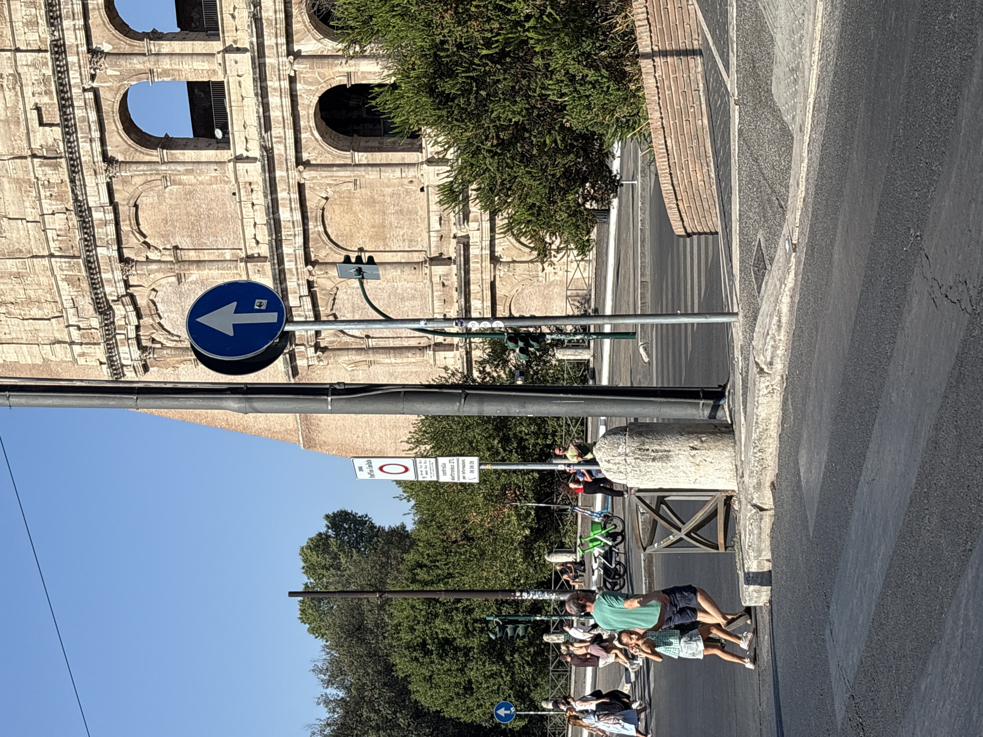 This image captures a sunny day scene in Rome, Italy, near the Colosseum. In the foreground, a pedestrian crossing is visible on a paved road. To the left, a group of people, including a man in a green shirt and a young girl in a plaid dress, are crossing the street. The man is gesturing with his hand, and the girl appears to be looking towards the camera. In the background, the iconic Colosseum's ancient brick structure dominates the scene, with its arched openings and weathered stonework. A traffic light is mounted on a pole, and a blue circular sign with a white arrow pointing upwards indicates a one-way street. Several trees and bushes line the area, and in the middle ground, a few bicycles are parked. The sky is clear and blue.