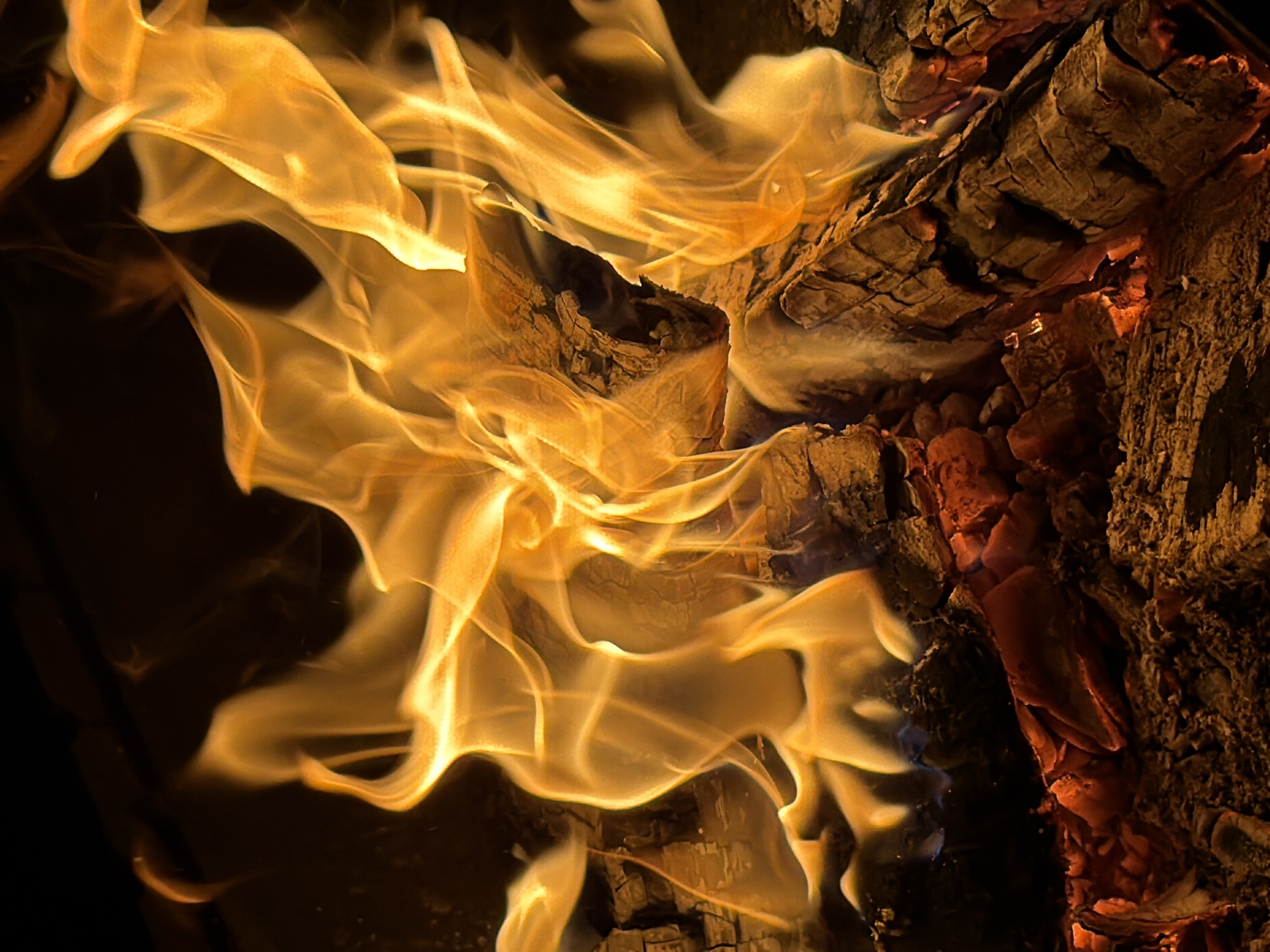 The image shows a close-up view of a fire burning. Flames in shades of yellow and orange engulf pieces of wood, whose surface is dark and cracked. The fire appears to be contained, suggesting it is in a fireplace or controlled burning area. The black areas surrounding the fire suggest a darkened environment. There are no people visible in the image. The location in Zimyonki, Russia is not evident from the image.
