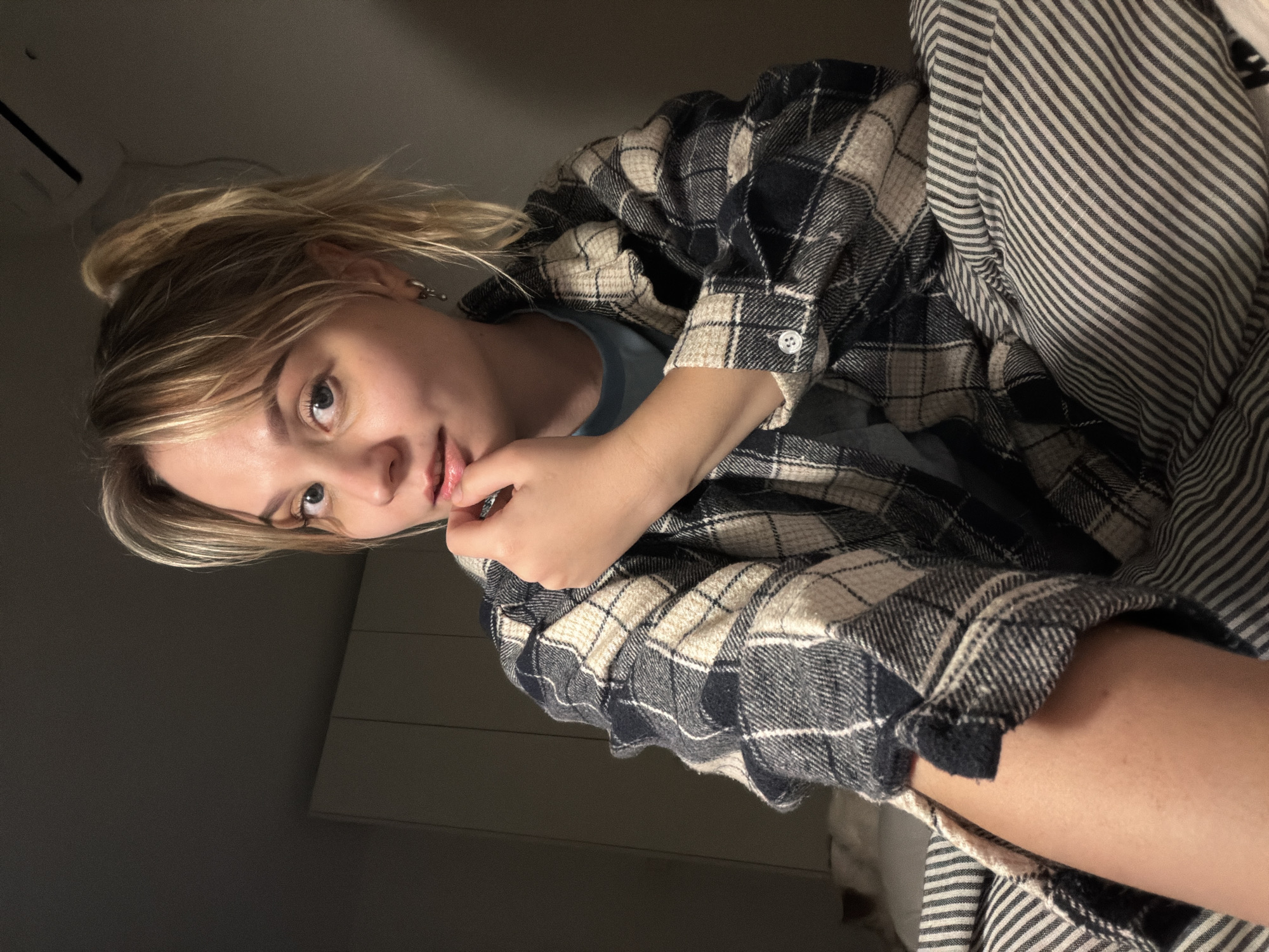 A young adult female with blonde hair tied in a high ponytail, with bangs falling across her forehead, and blue eyes, is depicted indoors, likely in a bedroom in Tel-Aviv, Israel. She is wearing a blue and beige plaid button-up shirt over a light blue t-shirt. Her right hand is raised, with her fingers lightly touching her chin and lower lip. Her left arm is visible in the lower left portion of the frame. She is looking directly at the camera. She is seated on a bed, partially covered by a white and dark grey striped duvet or blanket that extends across the lower and left side of the image. In the background, on the left, are light-colored, possibly white, built-in cabinets with vertical seams. The wall behind her to the right is a plain dark grey, with a portion of an air conditioning unit or ventilation grill visible in the upper right corner. The scene is illuminated by dim, ambient light.
