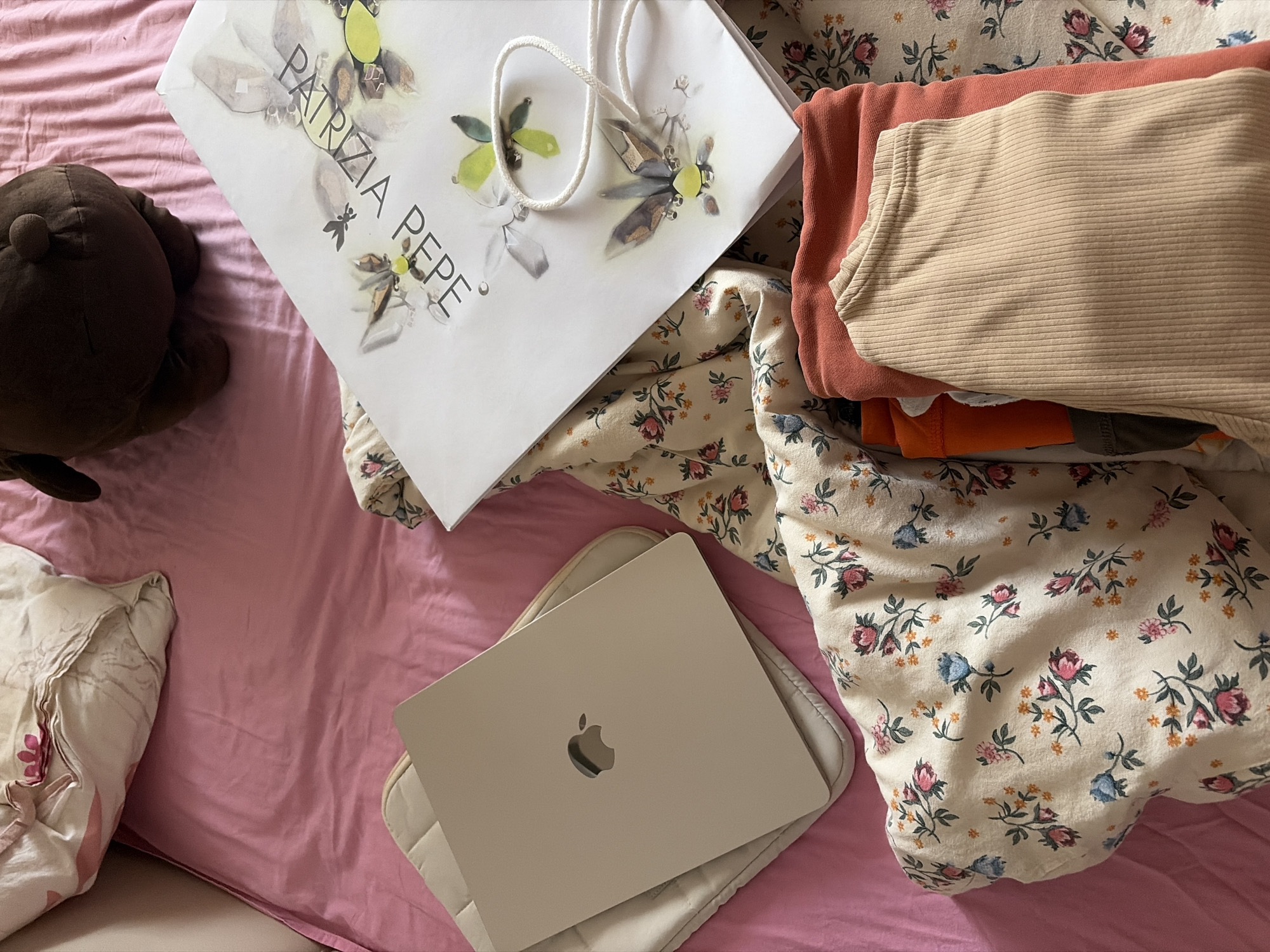 The image depicts a domestic bedroom scene. A pink comforter covers the bed, upon which rests a light-colored laptop, an Apple device, positioned on top of a quilted pad. Adjacent to the laptop is a shopping bag with the text "PATRIZIA PEPE" and floral imagery. A stuffed brown bear toy is situated at the top of the image. In the foreground, a stack of folded clothing items, including an orange t-shirt and a beige ribbed top, is visible on a floral patterned fabric. The overall setting appears to be a bedroom. The provided information does not specify a location in Burgas, Bulgaria.
