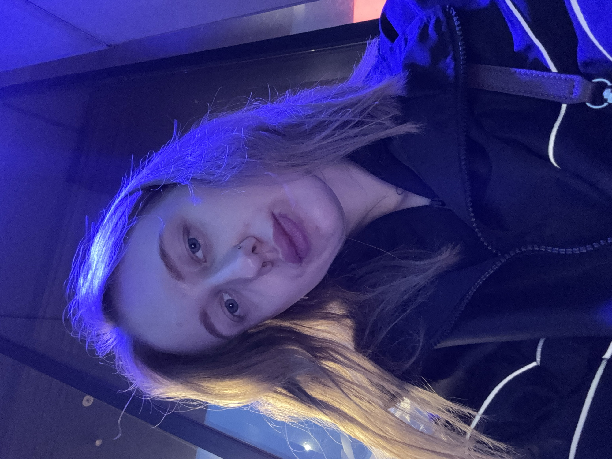 A young woman with long, light brown hair is photographed indoors, likely in Ol'ginskaya, Russia, given the prompt. Her hair is lit with a blue cast at the top of her head, fading into a more natural brown color towards the ends. She is wearing a dark jacket with a zipper and white stripes. Her face is visible, showing light skin, blue eyes, and a piercing in her left nostril. The background is dark, possibly a wall or window at night. The photo is taken from a slightly elevated angle.