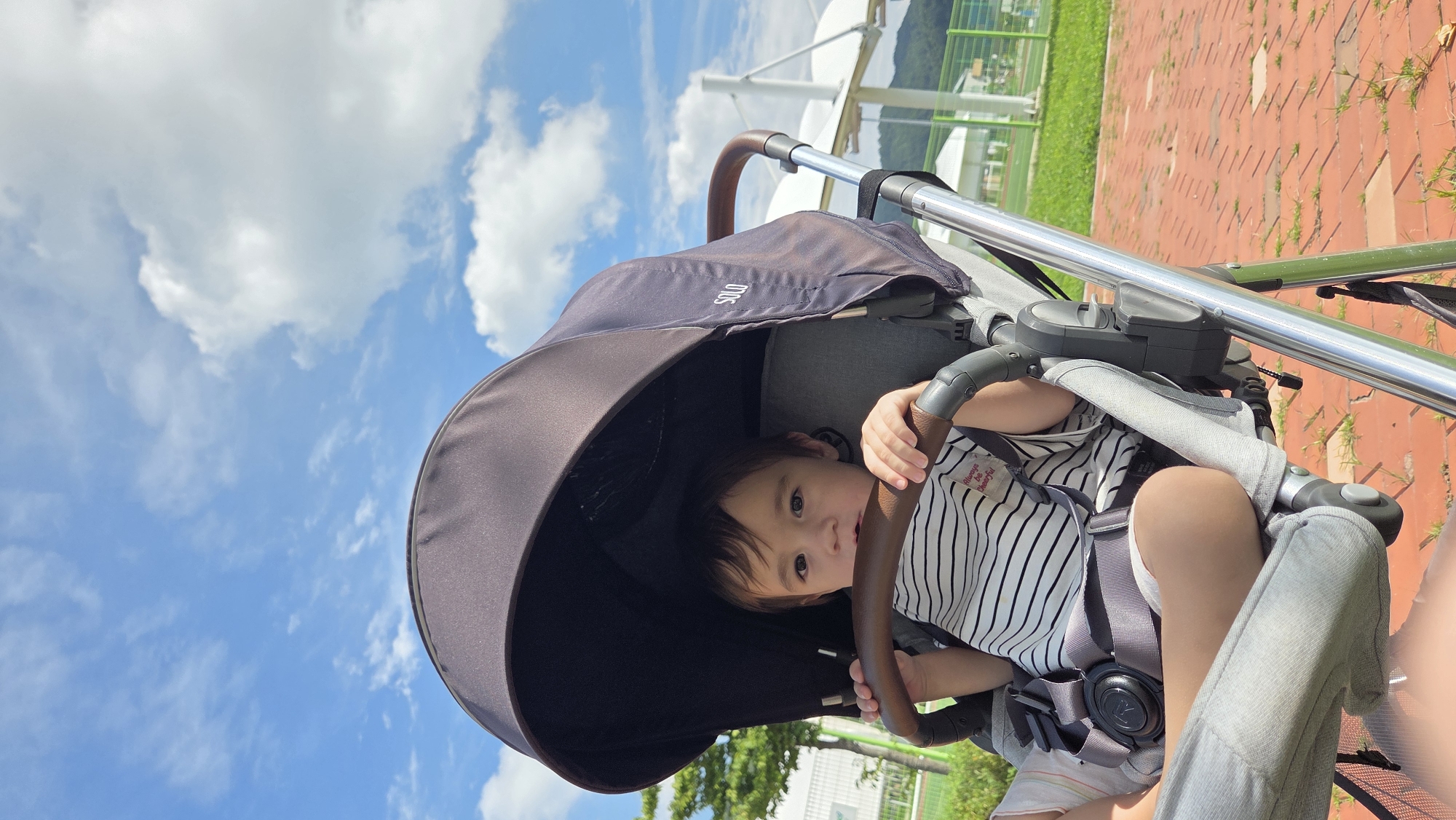 A young child is seated in a stroller outdoors under a blue sky with white clouds. The child is wearing a black and white striped t-shirt and light-colored pants, with a gray safety harness secured around them. The stroller has a dark canopy providing shade and a brown handlebar. The background shows a paved area with red bricks, green grass, trees, and a white structure that appears to be part of a park or recreational facility. The location is identified as Dunnae-myeon, South Korea.
