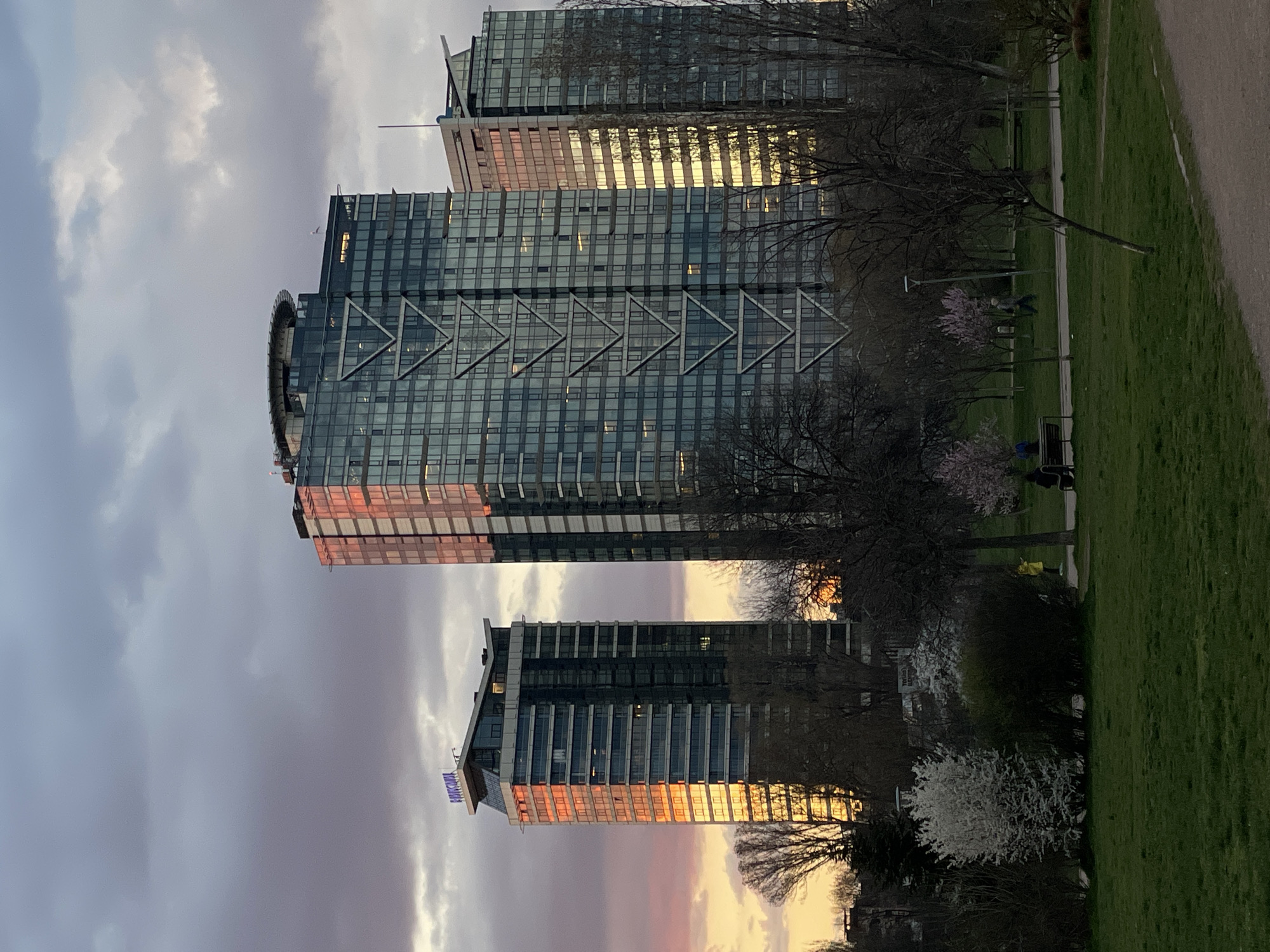Captured from a low angle, the image showcases a park environment in Sofia, Bulgaria, with three contemporary high-rise buildings in the background. The buildings, reflecting the warm hues of the setting sun, add a touch of modernity to the natural surroundings. A group of people are present in the park, adding life to the scene. The sky is filled with clouds illuminated by a sunset, giving a vibrant contrast to the otherwise subdued tones of the image. Bare trees interspersed with those in full bloom, suggest the transition from winter to spring, enhancing the overall aesthetic of the landscape.