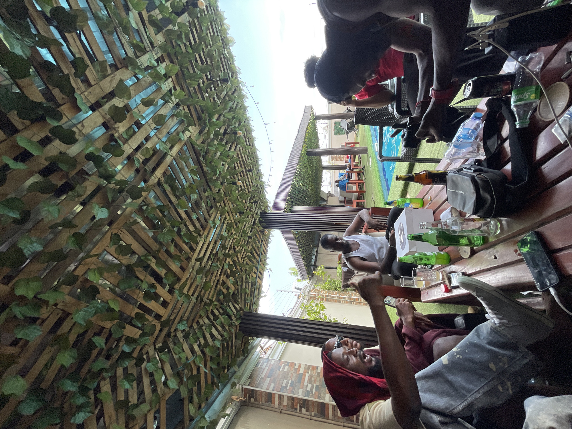 This image captures a casual outdoor gathering in Abuja, Nigeria, under bright daylight. The scene is set on what appears to be a patio or rooftop area, featuring a decorative wooden trellis adorned with artificial green foliage overhead, creating a canopy or wall feature. String lights are visible along the trellis.
Several young adults are present, relaxing and socializing. In the foreground, a man with a beard and a red head covering is prominently featured, looking upwards with a broad smile or laugh, holding a drink. His left arm is raised, suggesting excitement or a celebratory gesture. Another person wearing sunglasses is partially visible next to him, also smiling. Further back, another individual in a white tank top is seated, and additional people are visible at the far end of a wooden slatted table.
The table is laden with various beverages and personal items. Numerous bottles are visible, including green glass bottles (possibly beer or soda), a brown bottle, a clear plastic Sprite bottle, and a plastic water bottle. Aluminum cans and glasses containing drinks are also present. Other items on the table include a black bag with "FASHION" written on it, a white box (possibly for a cake or gift), and a mobile phone (likely an iPhone) plugged into a charger.
The background shows additional outdoor elements, such as a tiled floor, green artificial turf, and distant structures, including another trellis-covered area and what might be the edge of a swimming pool. The clear, sunny sky indicates pleasant weather, and the overall mood conveyed by the subjects appears to be joyful and relaxed.