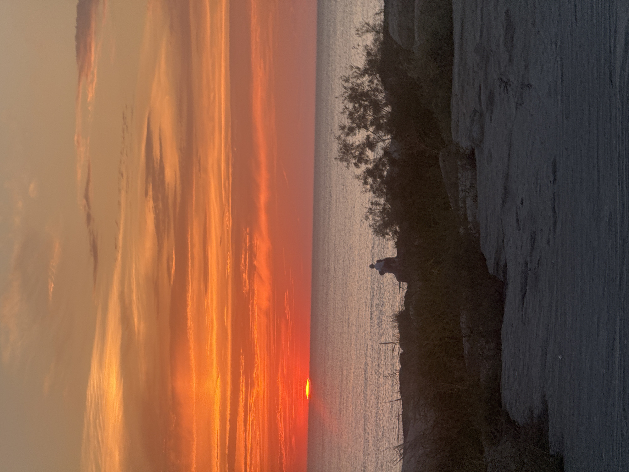 A sunset over the Caspian Sea is captured from a rocky shoreline, likely in Aktau, Kazakhstan. The sun, a bright orange orb, is partially submerged on the horizon, casting vibrant orange and yellow hues across the sky and reflecting on the calm water. Wispy clouds stretch horizontally, accentuating the sunset's colors. In the foreground, silhouetted figures, possibly a couple, sit together on a rocky outcrop, their forms dark against the illuminated sea and sky. Vegetation, including dry grasses and bushes, grows amongst the rocks. The ground in the immediate foreground is sandy and uneven.